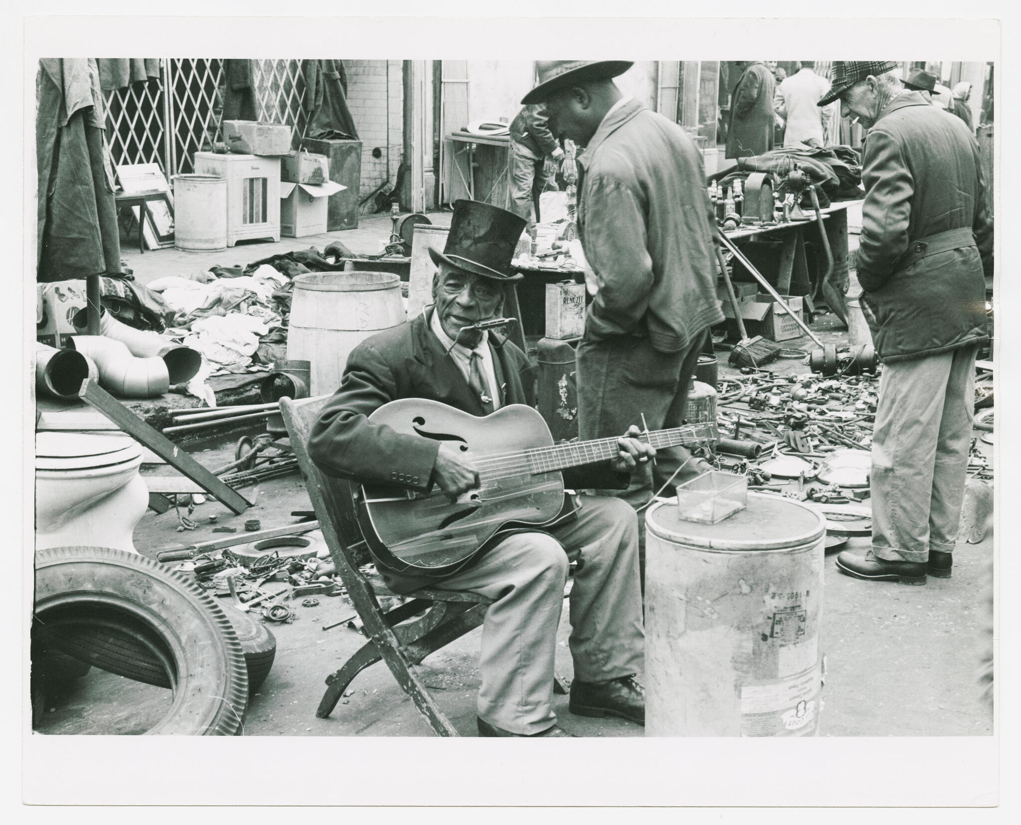 Man wearing a top hat plays guitar and harmonica while seated amid junk and market stalls.