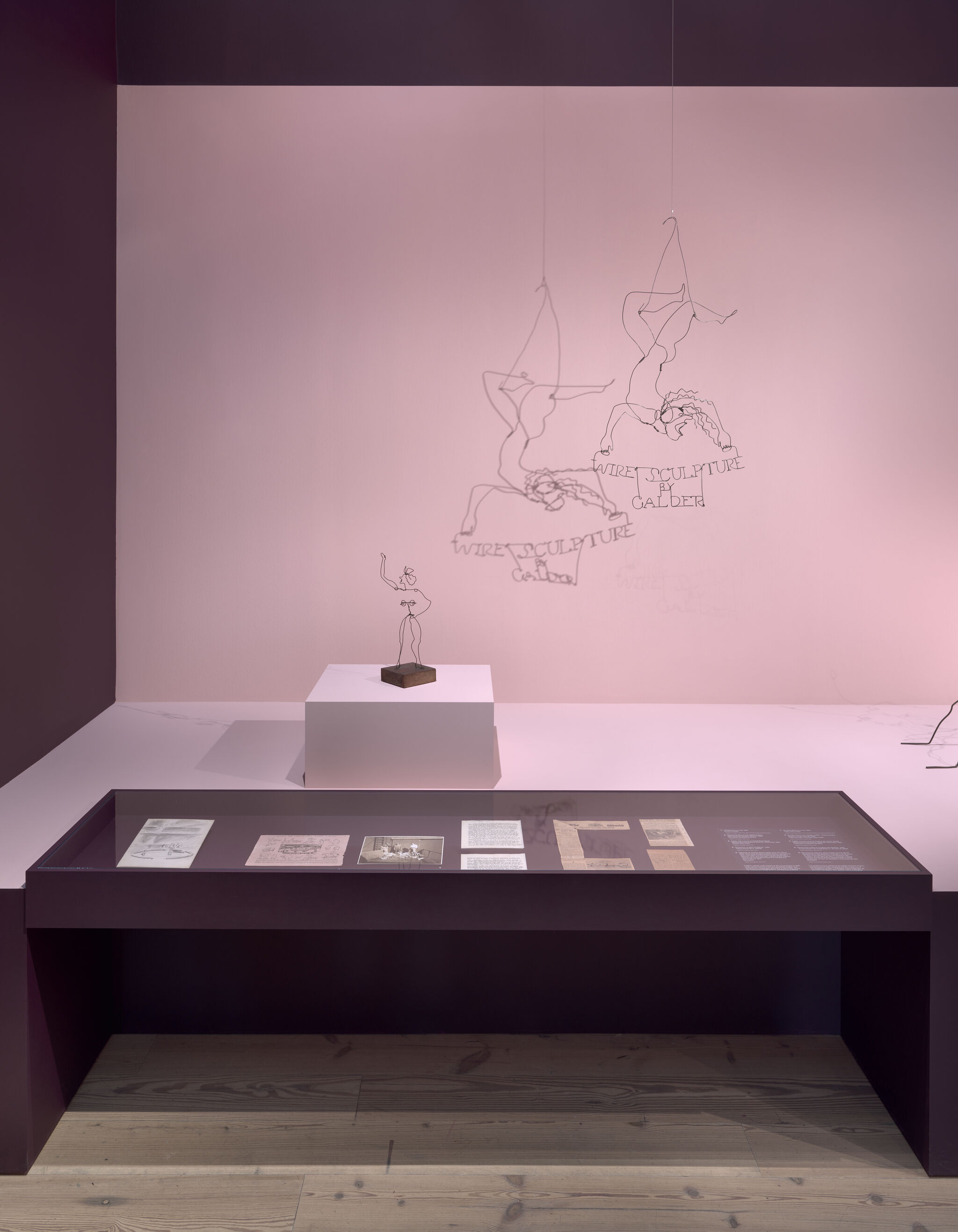 Wire sculptures hang and stand above a pedestal and display case against a pink wall.