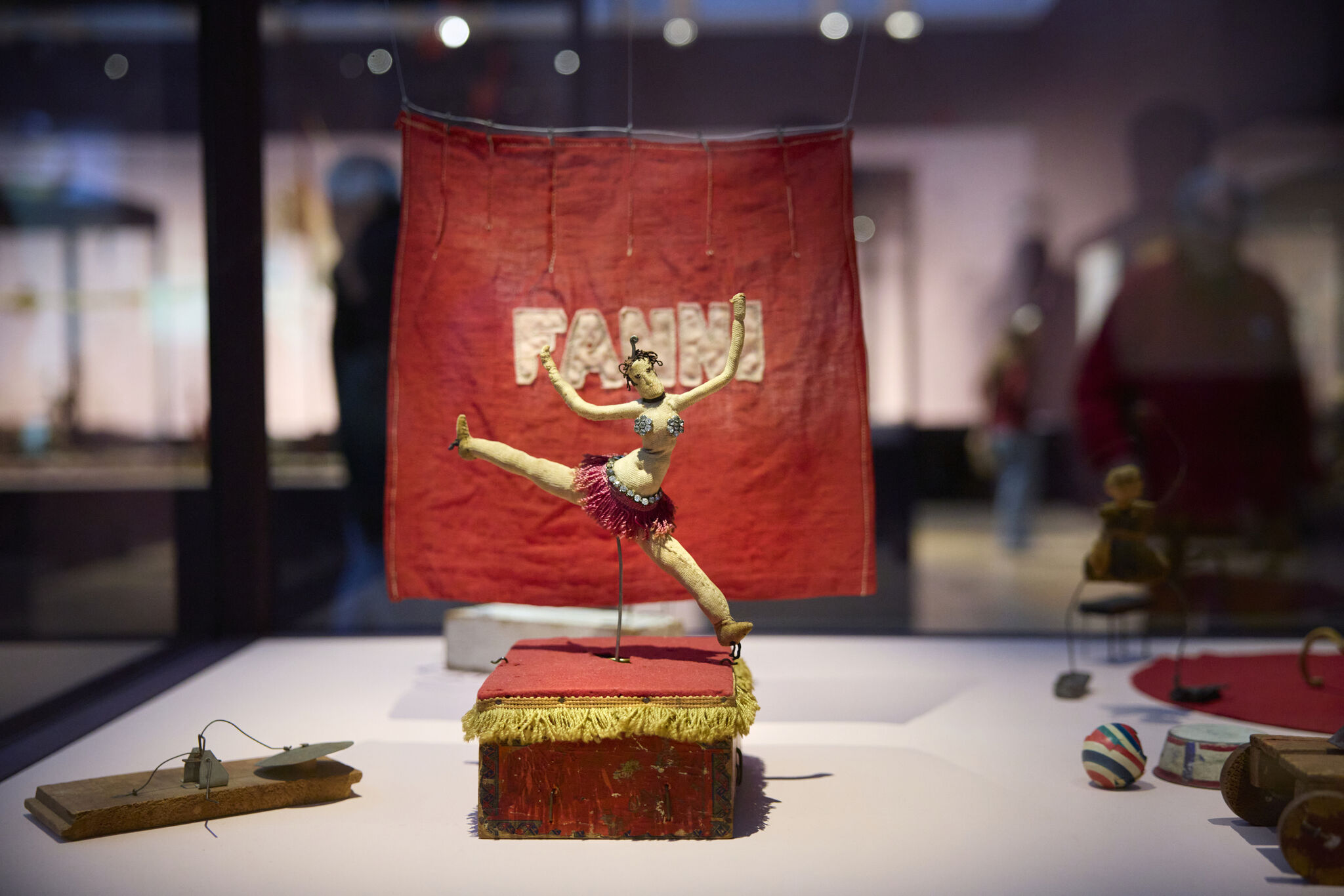 A small vintage circus dancer puppet in a leaping pose on a red fringe-covered platform.