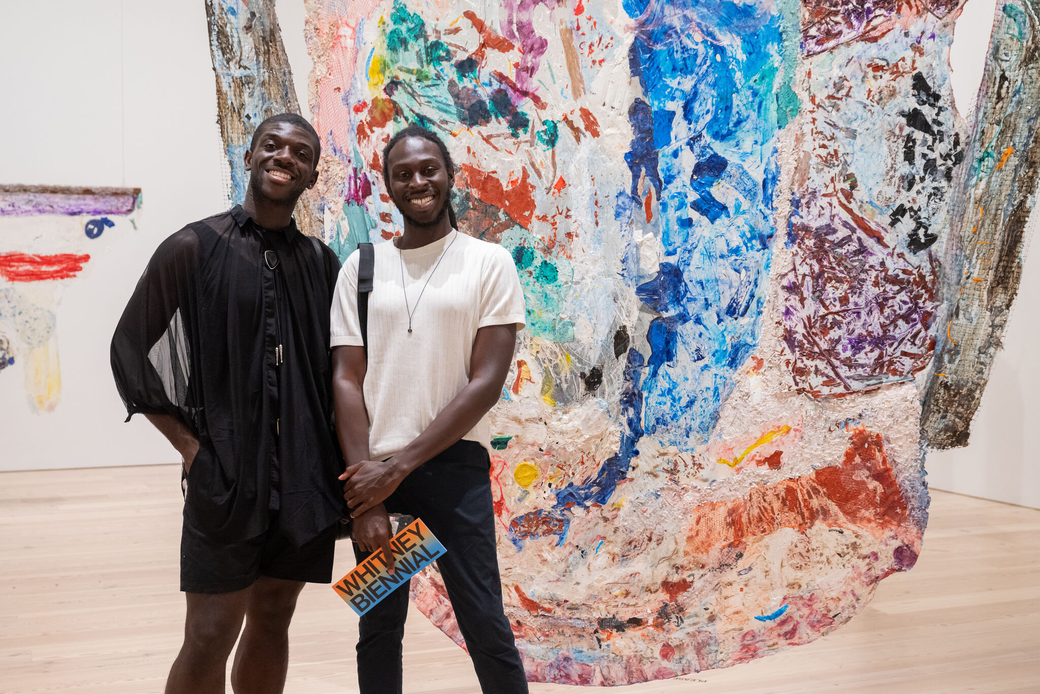 Two smiling men stand in front of a large colorful abstract artwork, one holding a Whitney Biennial pamphlet.
