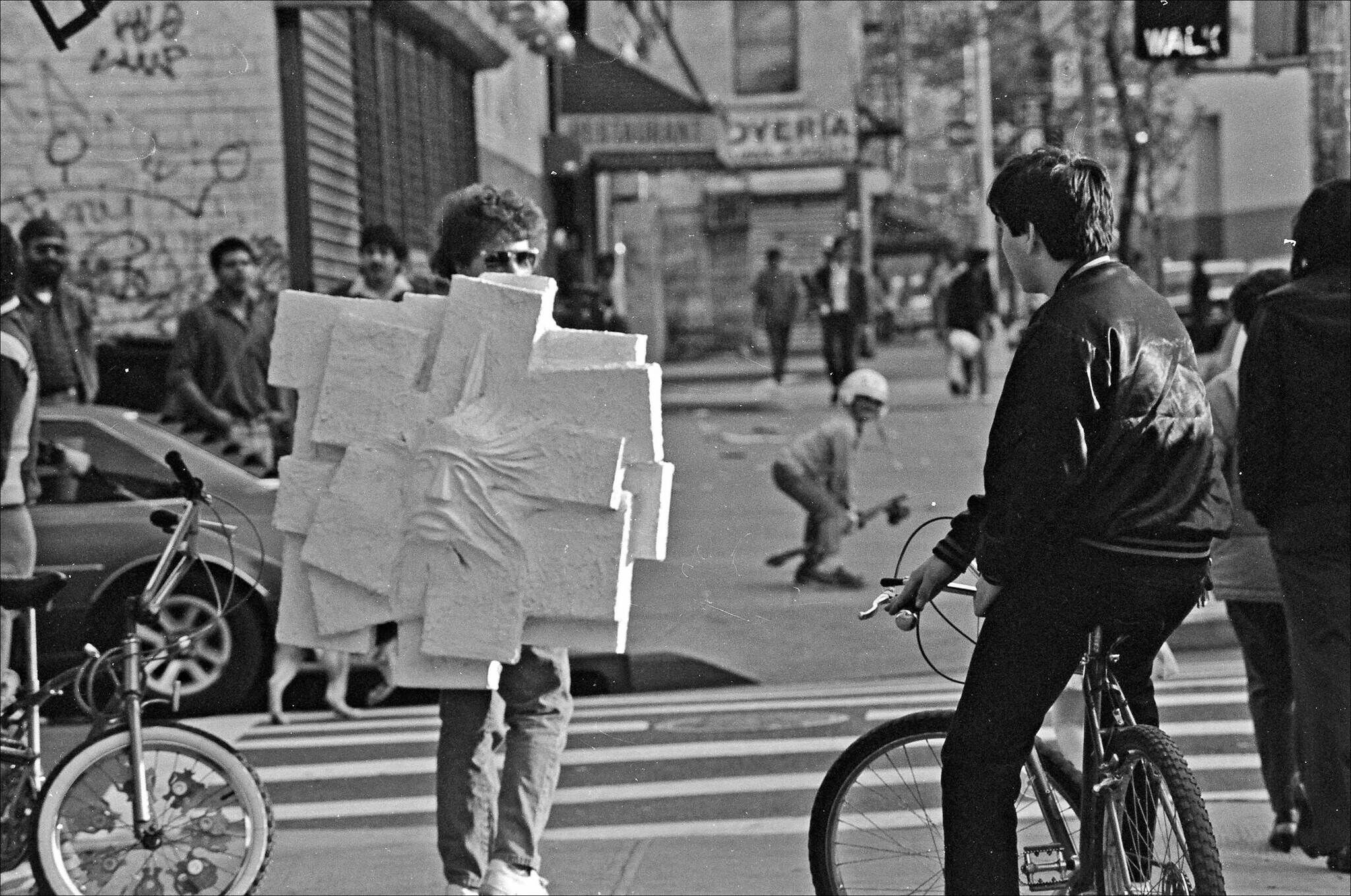 Person wearing a large foam sculpture of a face walks across a crosswalk toward a cyclist.