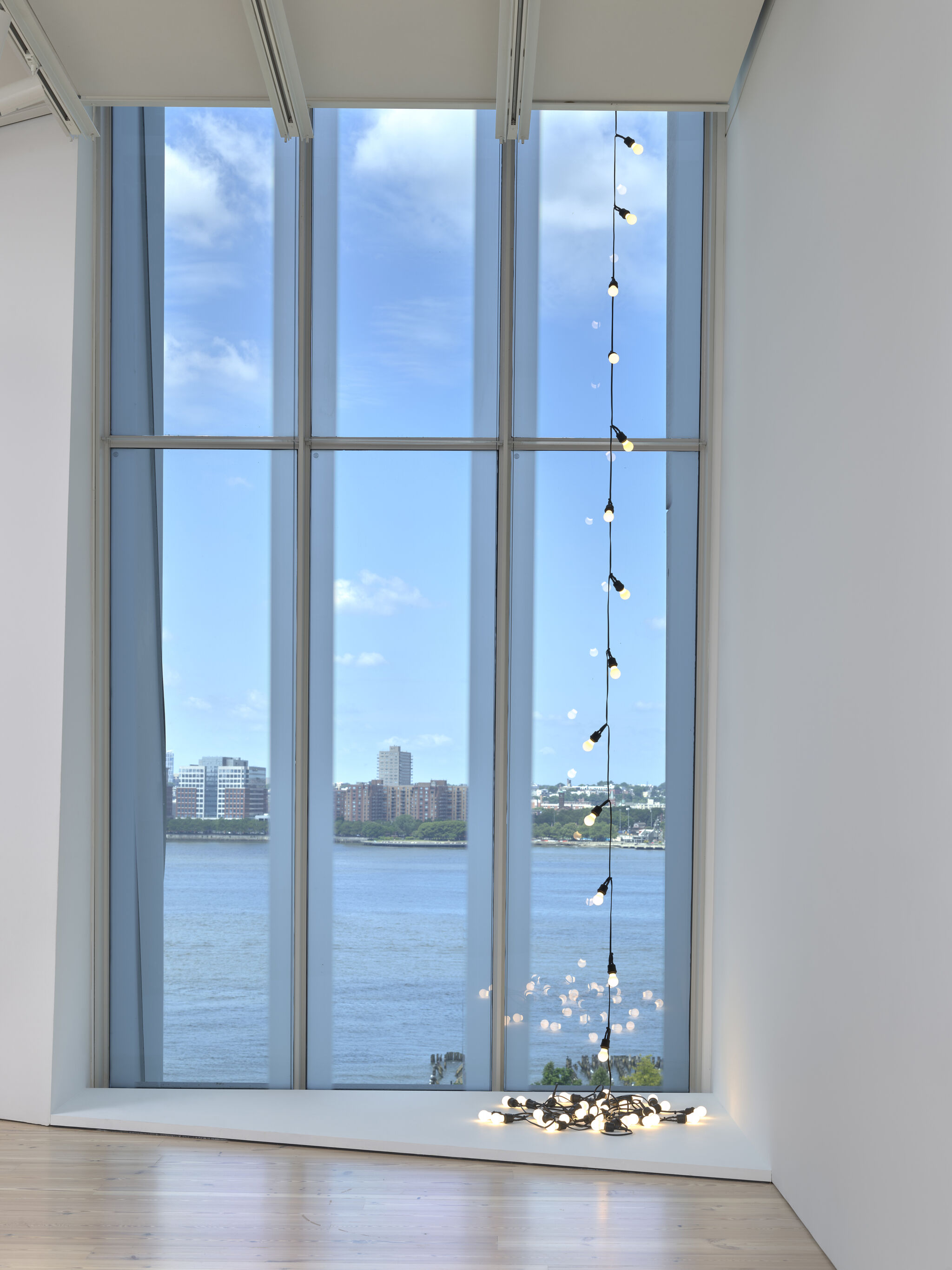 A string of hanging light bulbs cascades downward, with several bulbs lying on the floor in front of a window overlooking Hudson river. 