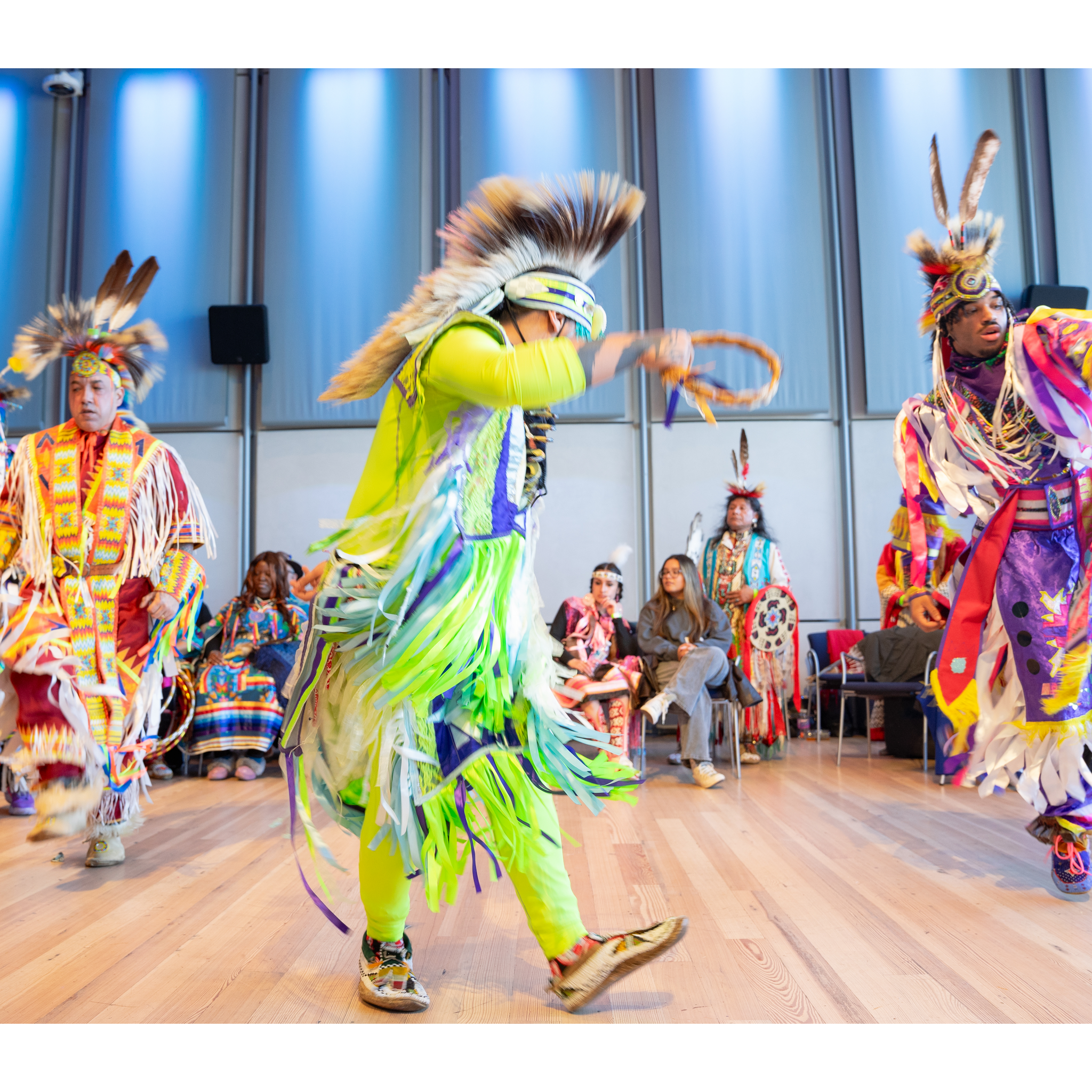 Four dancers in traditional dress actively dancing in front of a seated audience
