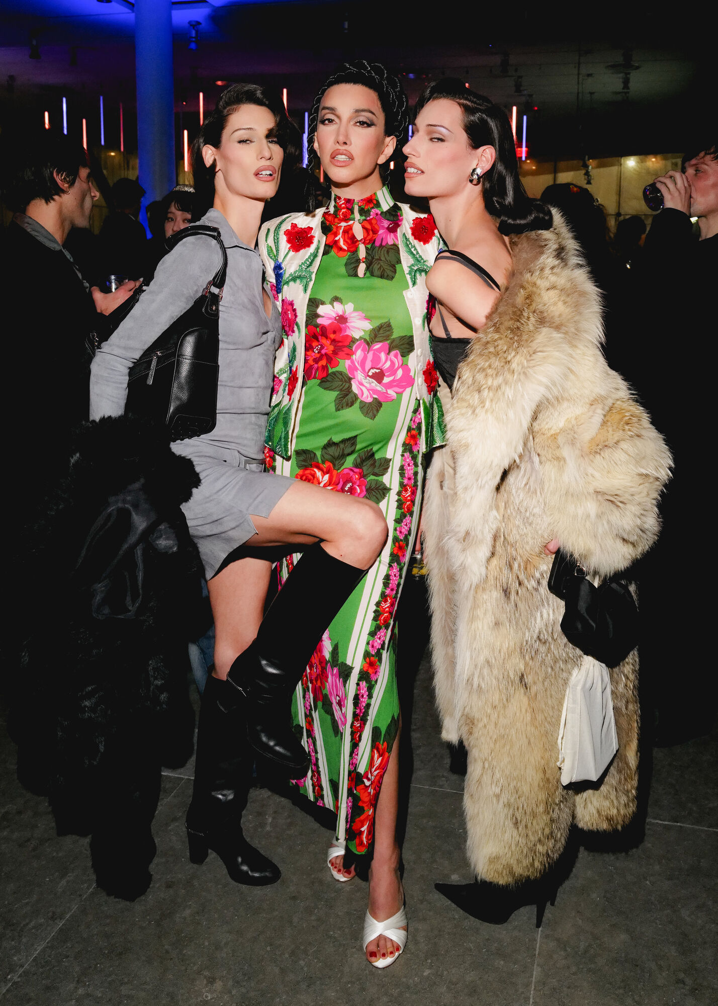 Three women pose closely together at a party, the center wearing a green floral dress.