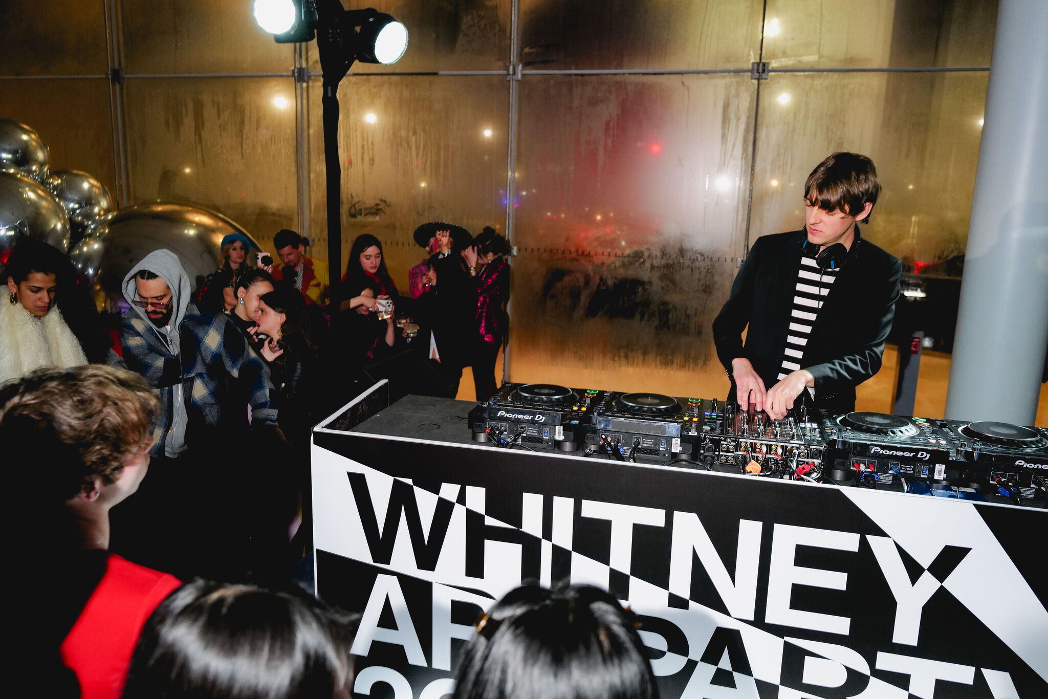 A DJ adjusts controls at a mixer while a crowd watches at Whitney Art Party.