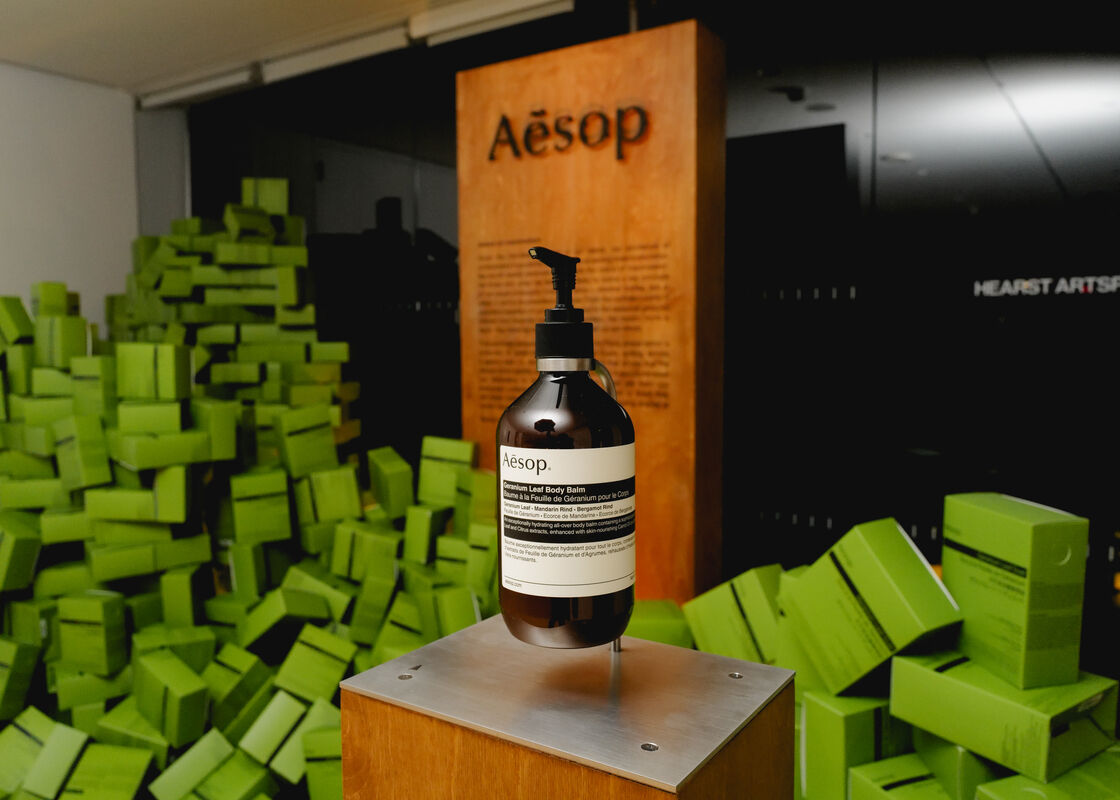 Aesop lotion bottle displayed on a metal pedestal with stacked green boxes behind.