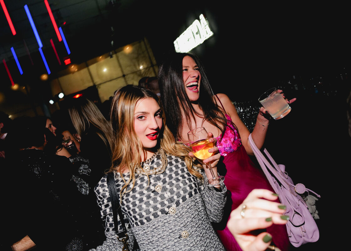 Two women at a crowded party holding drinks, one laughing loudly while posing for the camera.