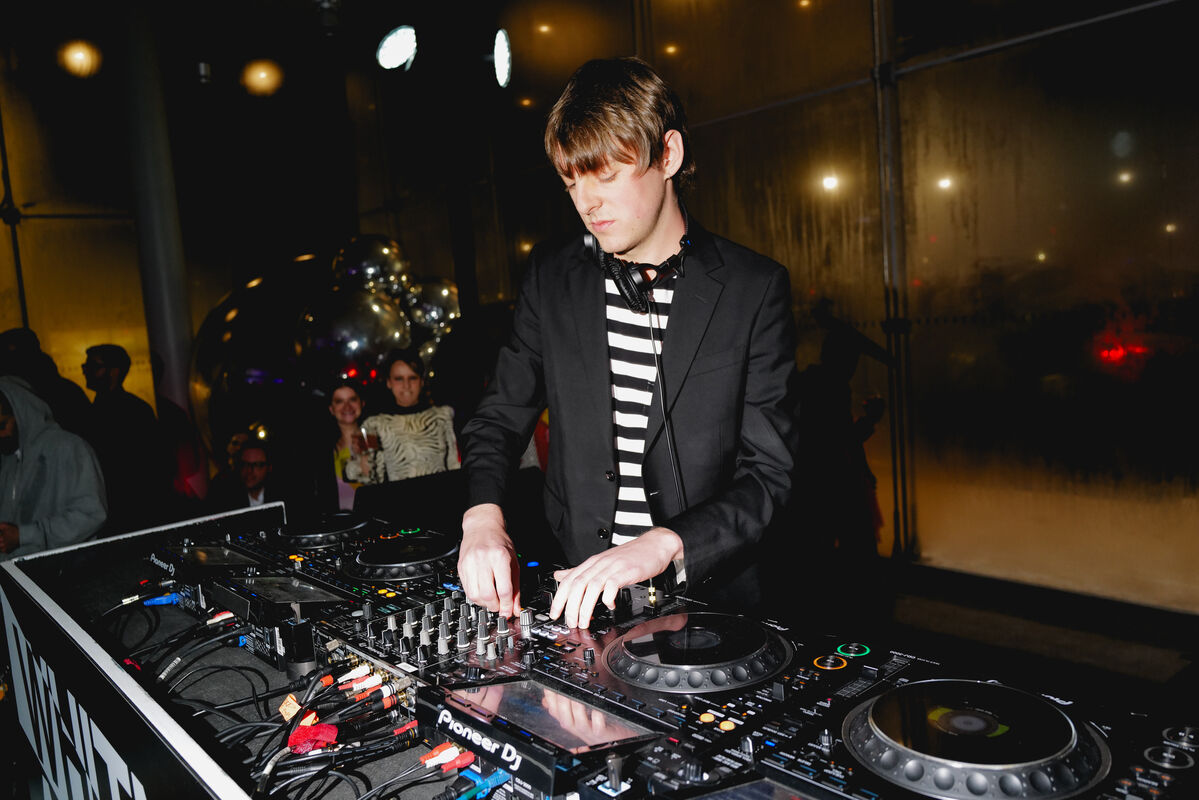 A DJ wearing headphones adjusts knobs on a mixer while people watch in the background.