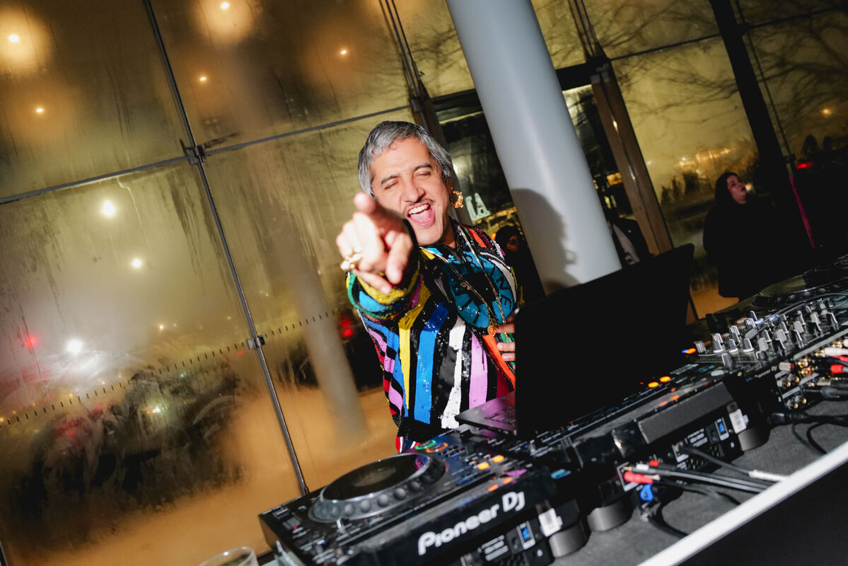 A smiling DJ in a colorful jacket points toward the camera while playing on a Pioneer DJ setup.