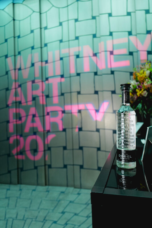 A bottle of Dobel tequila sits on a glossy table in front of a projected Whitney Art Party sign.