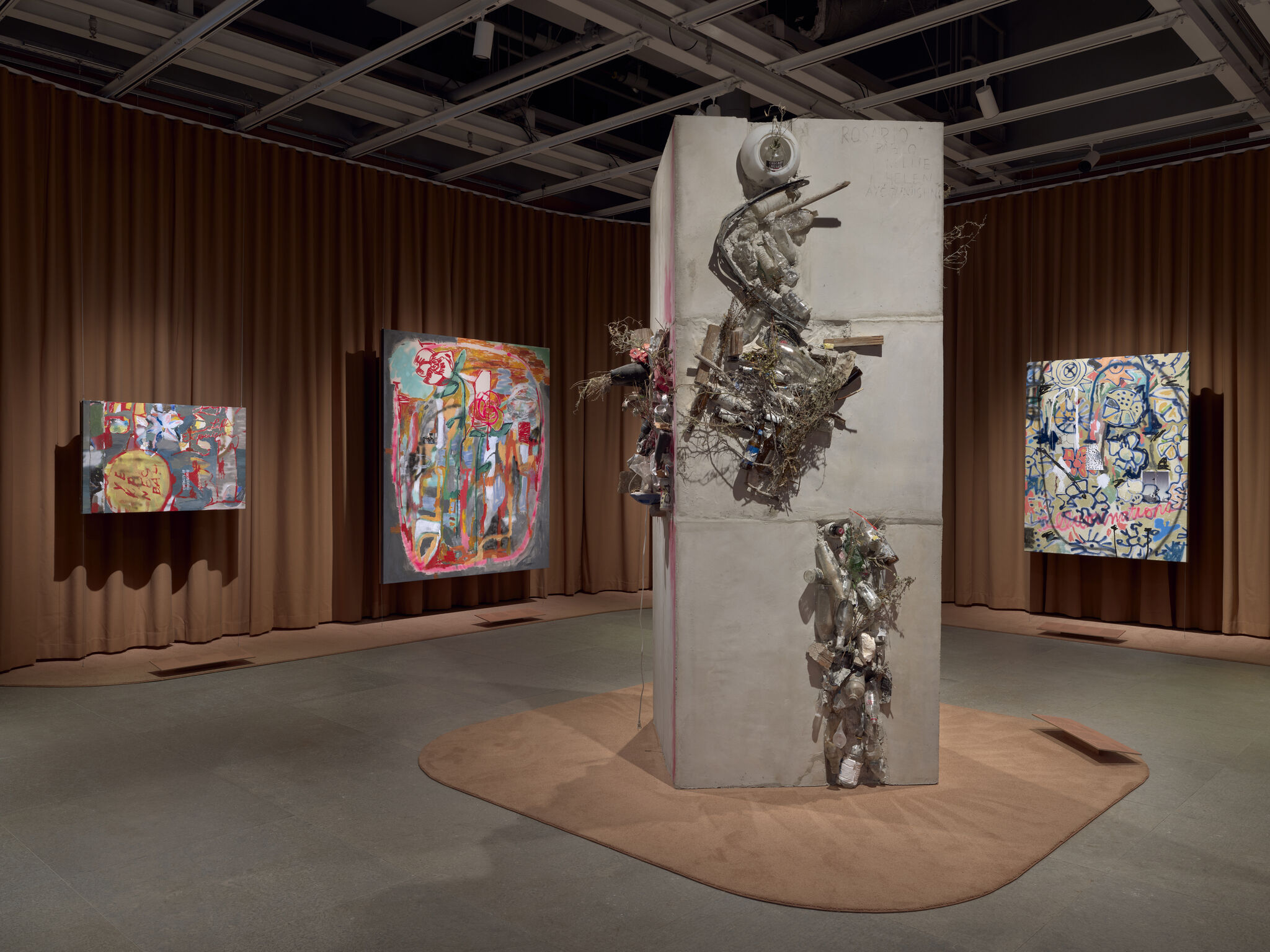 Central concrete pillar with mixed-media debris sculptures surrounded by colorful abstract paintings in a gallery.