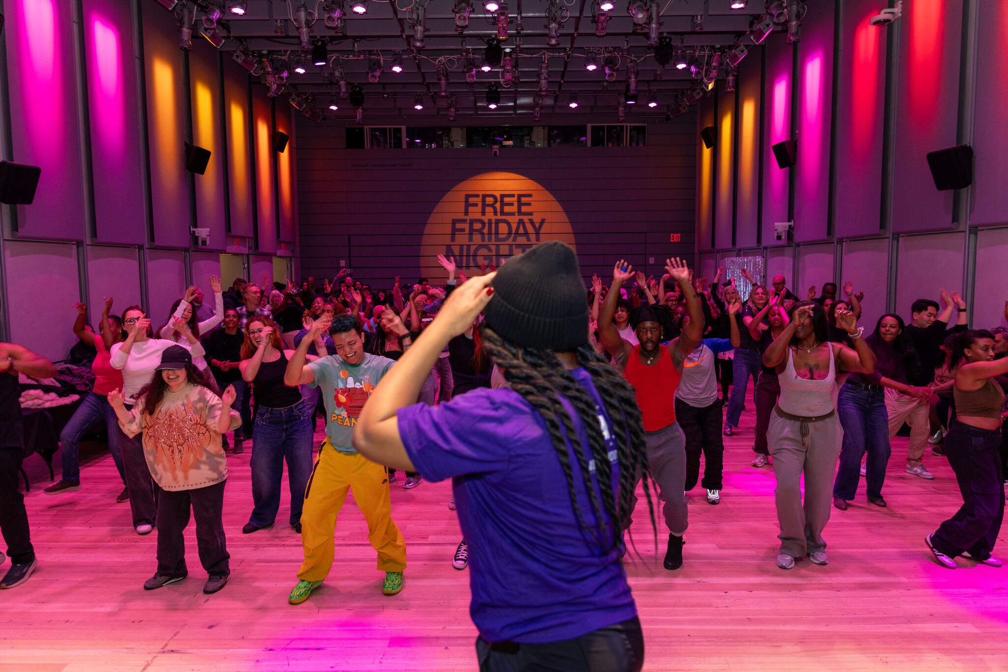Instructor leads a large group dancing in a colorful studio with "Free Friday Night" projected on wall.