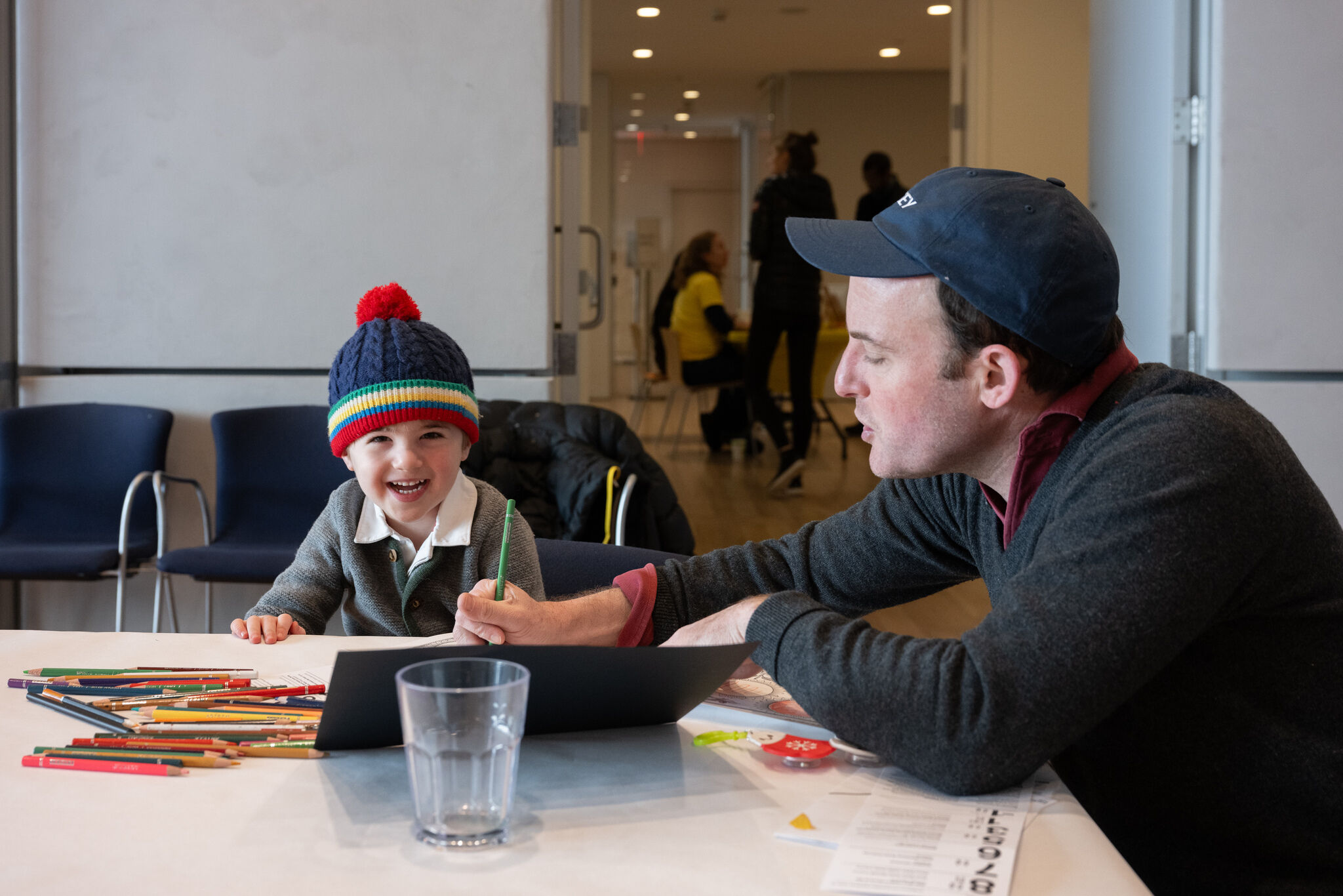 A smiling child in a knit hat watches as an adult draws with colored pencils.
