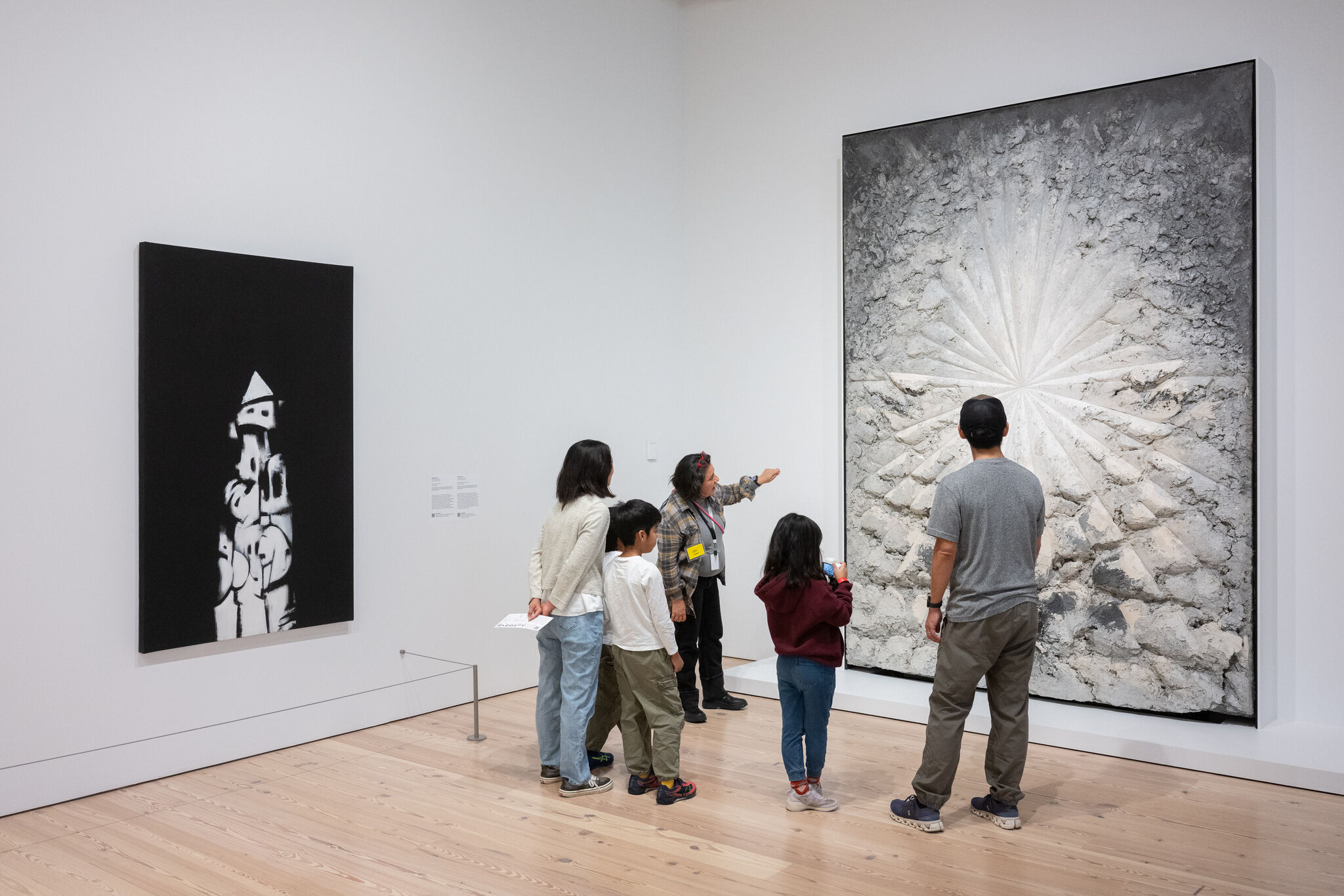 A museum guide points to a large textured wall artwork while a family observes closely.