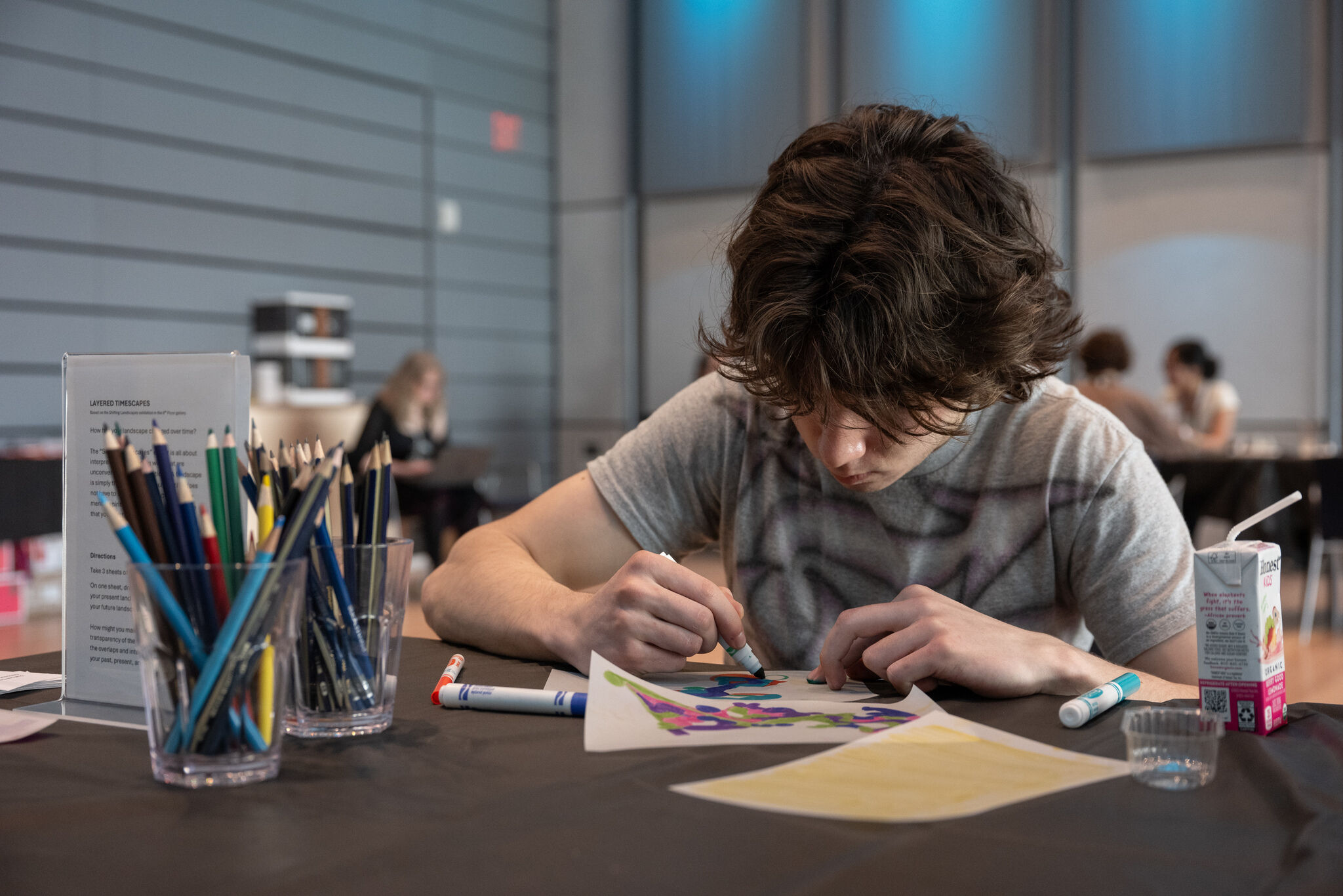 Teenager concentrates while coloring a colorful abstract drawing at a table with markers.