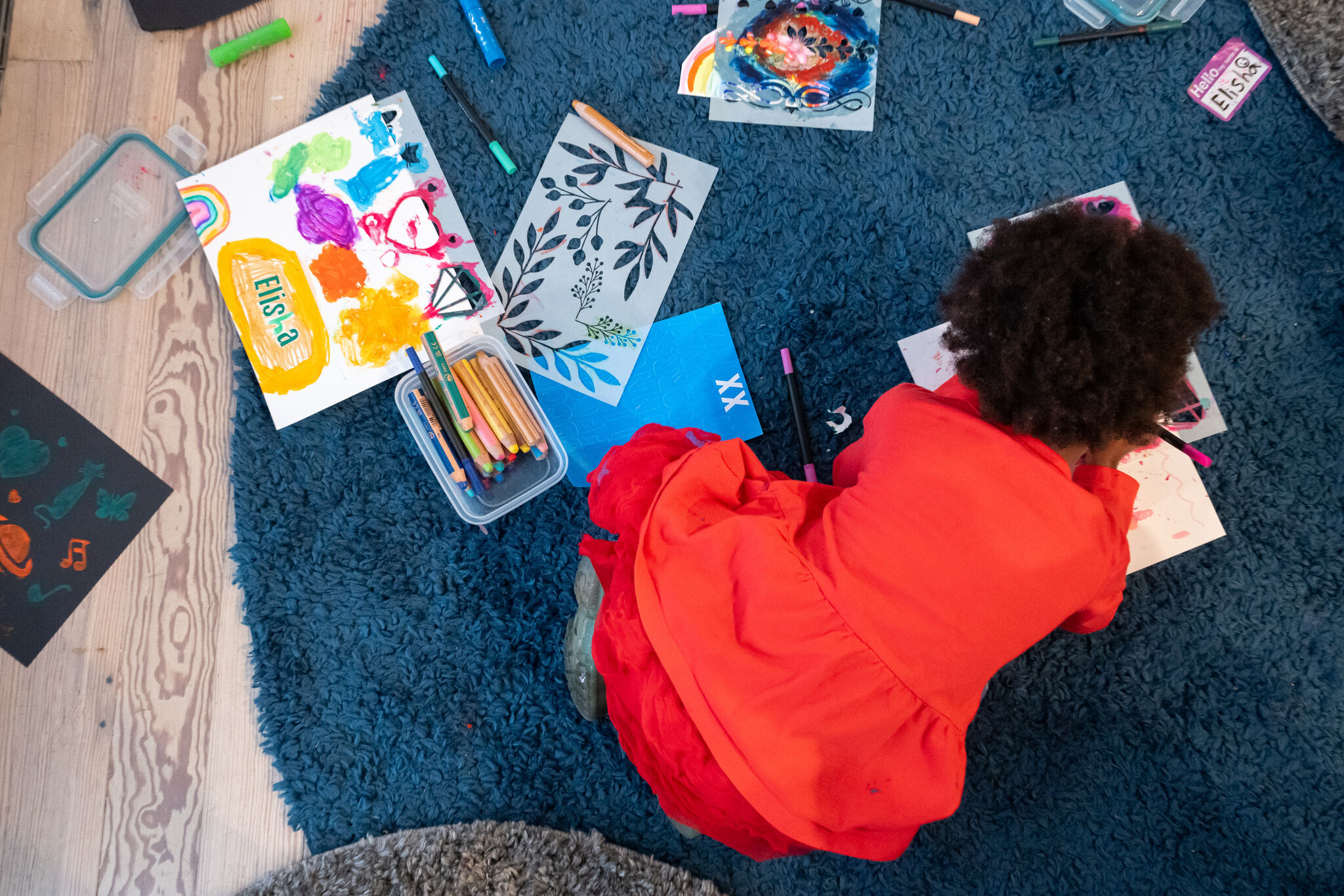 Child in a red dress lying on a blue rug drawing and coloring with scattered art supplies.