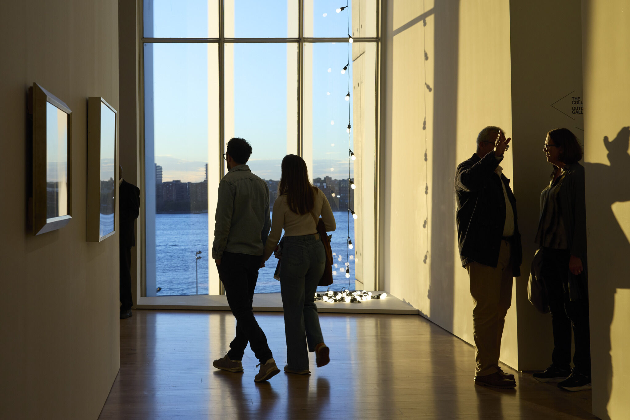 A couple walks hand in hand toward a large window overlooking the river at sunset.