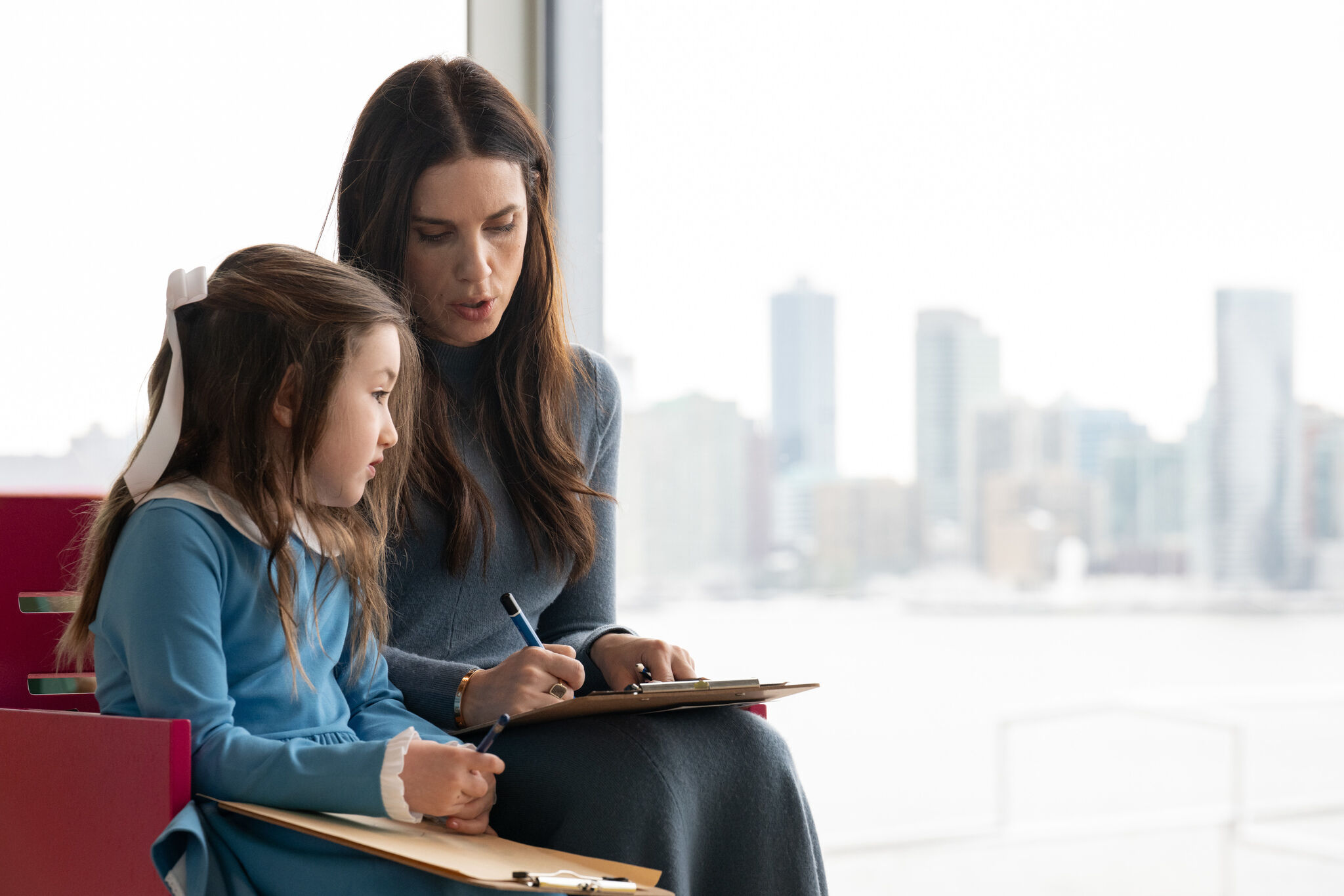 Woman helps young girl write on a clipboard while they sit by a window with city skyline.