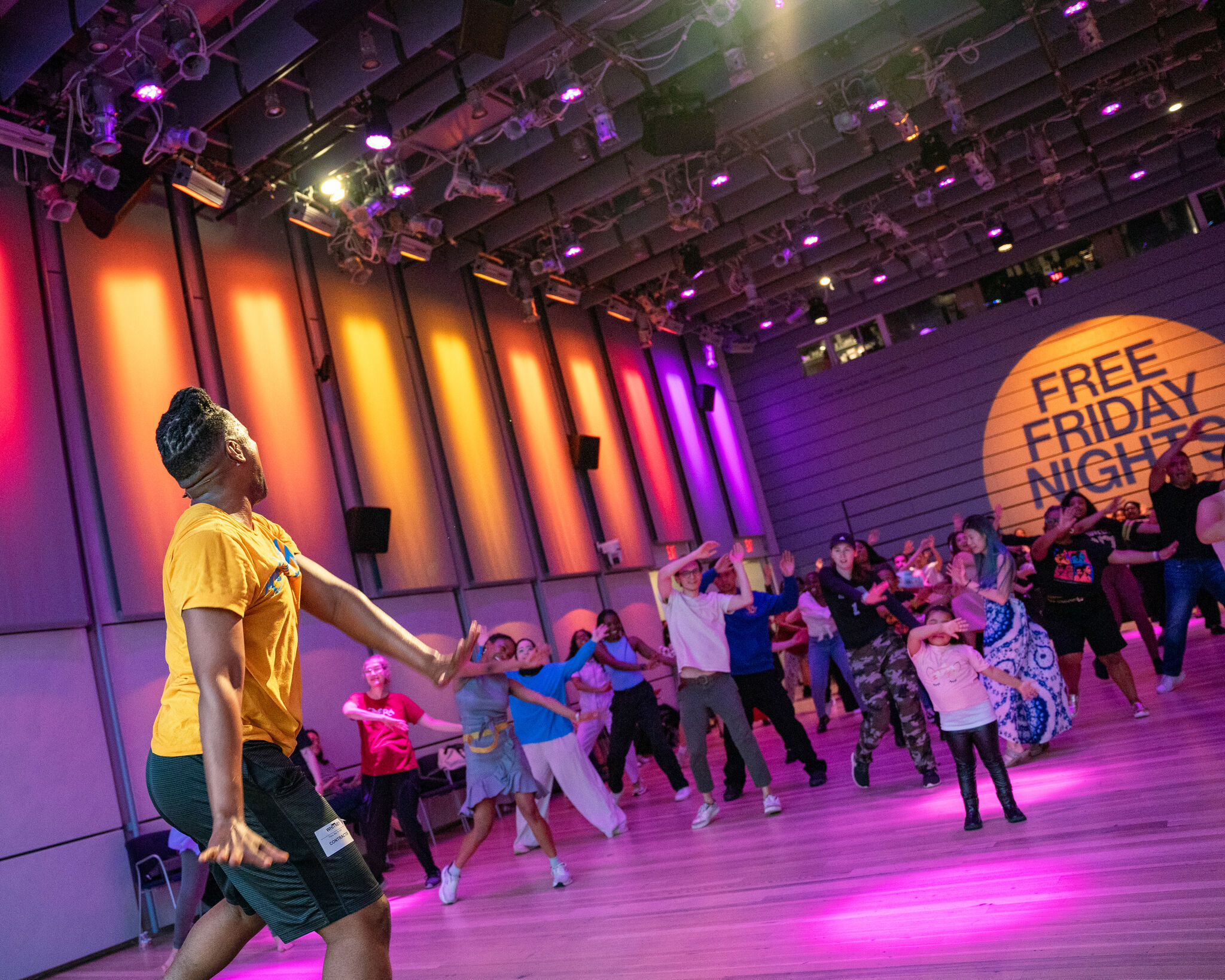 A dance instructor leads a diverse group in energetic moves under colorful lights with "Free Friday Nights" projected.