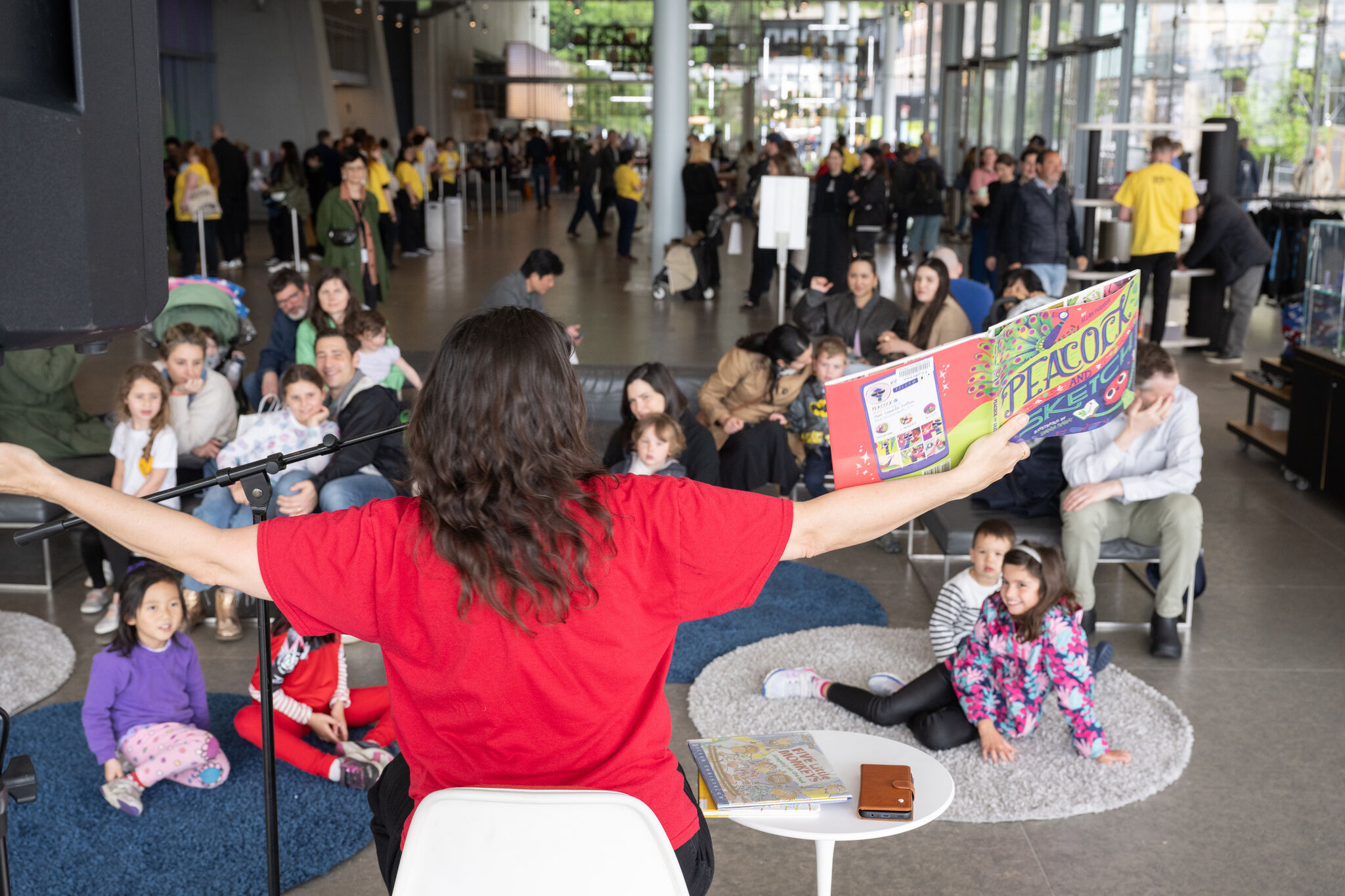 A storyteller reads animatedly from a colorful book to a group of children seated on rugs.