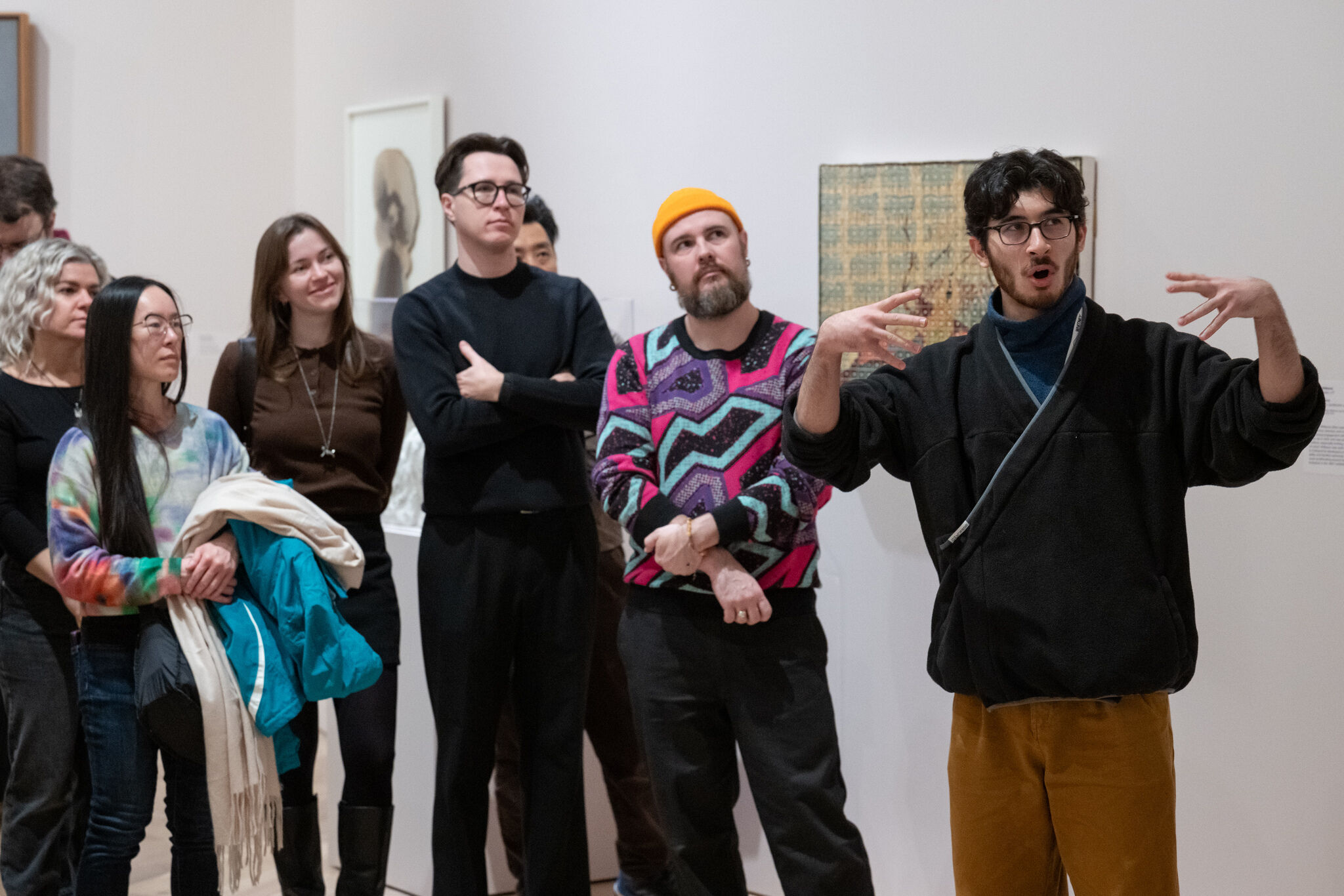 A young man gestures animatedly while leading a group tour in an art gallery.