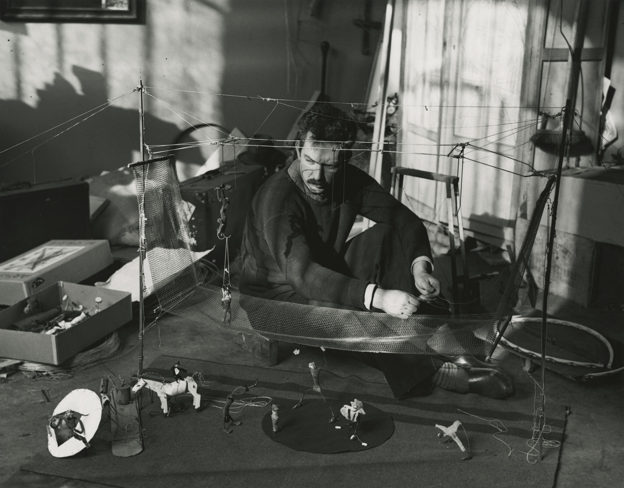 A man sits on the floor repairing a wire-and-mesh puppet stage with small figurines.