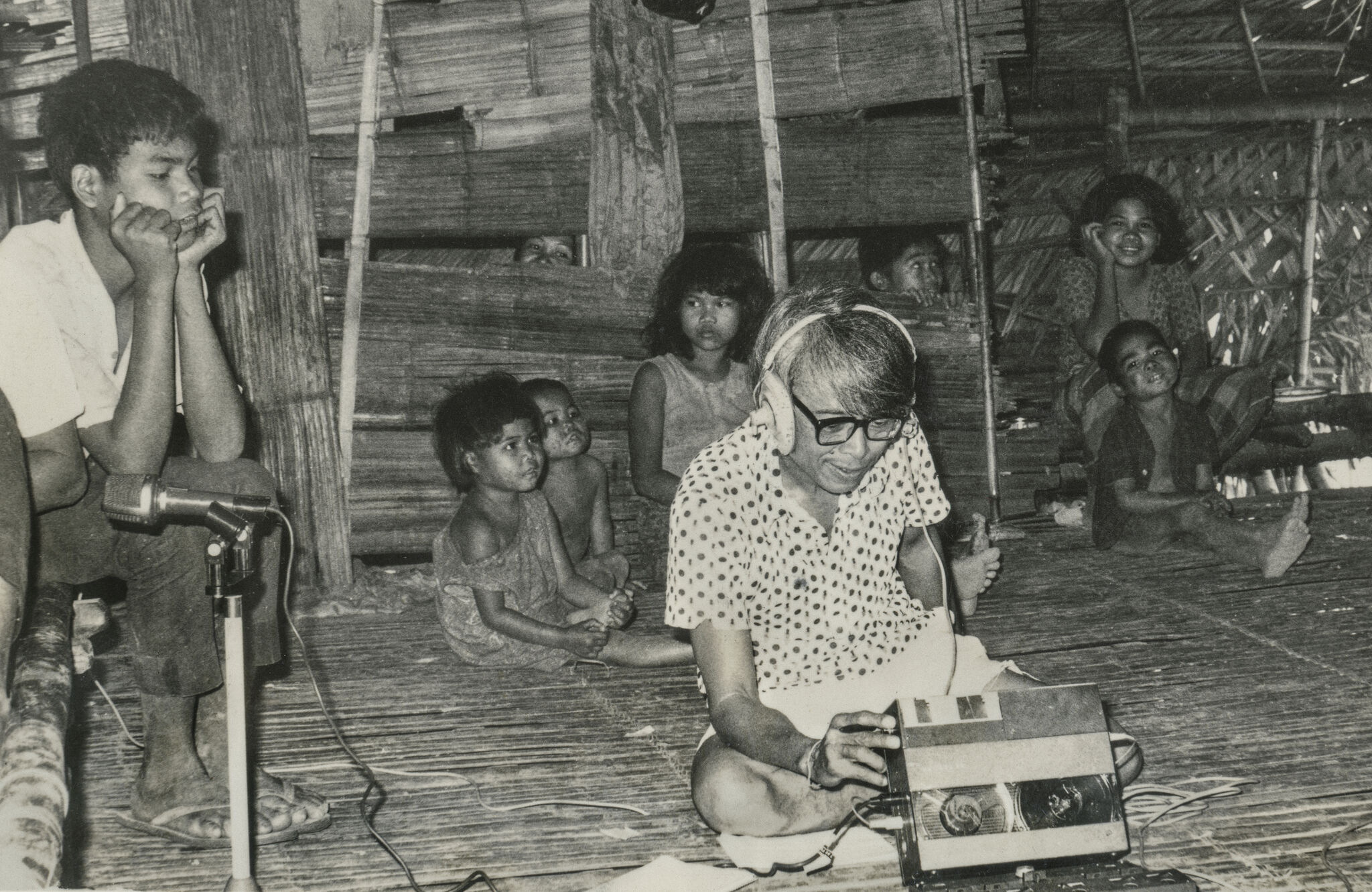 An elderly man wearing headphones operates a tape recorder while children watch inside a bamboo hut.
