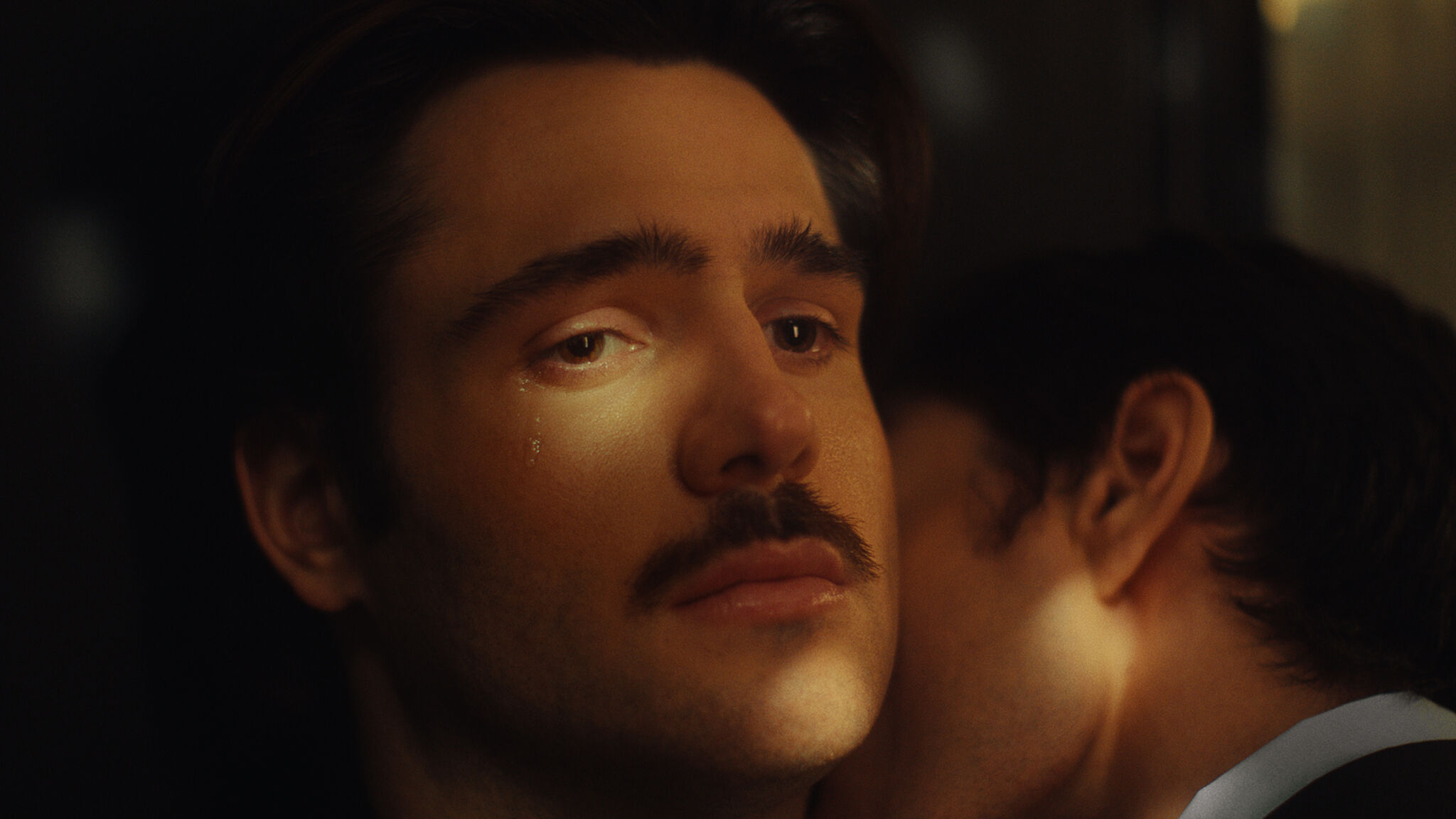 A man with a mustache has a tear on his cheek while someone whispers into his neck.