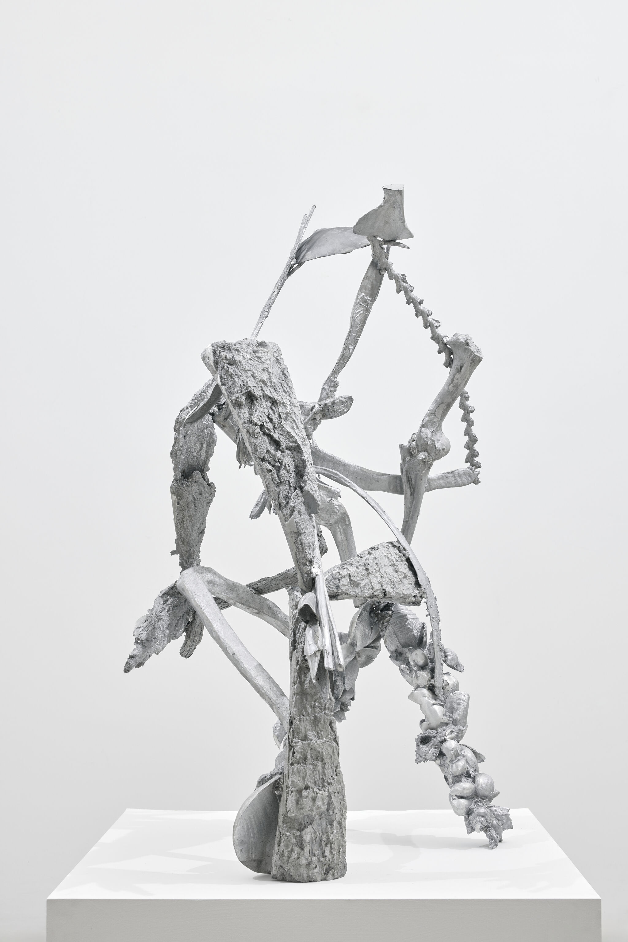 Abstract assemblage of intertwined weathered wood and organic forms painted gray on a white pedestal.