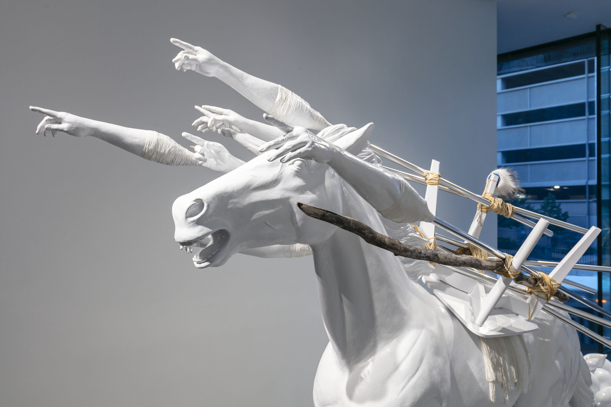 A white horse sculpture with multiple outstretched human arms pointing forward and wooden poles tied to its back.