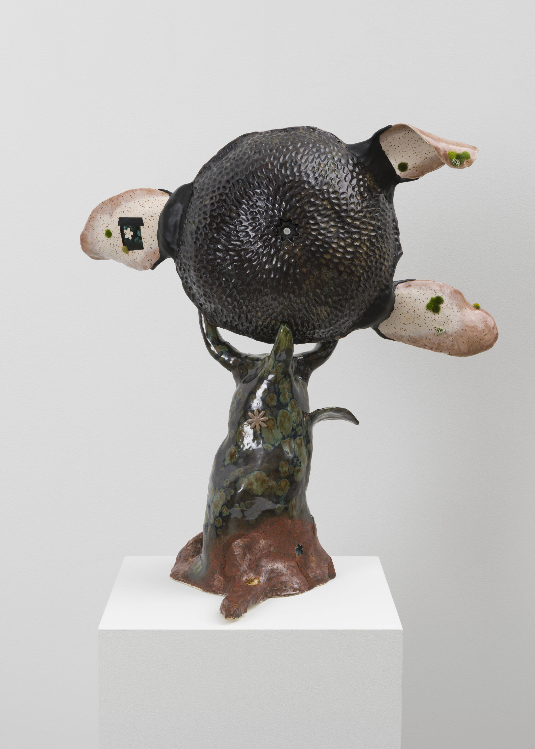 A glazed ceramic figure resembling a tree holds a large textured black disk with three pink appendages.