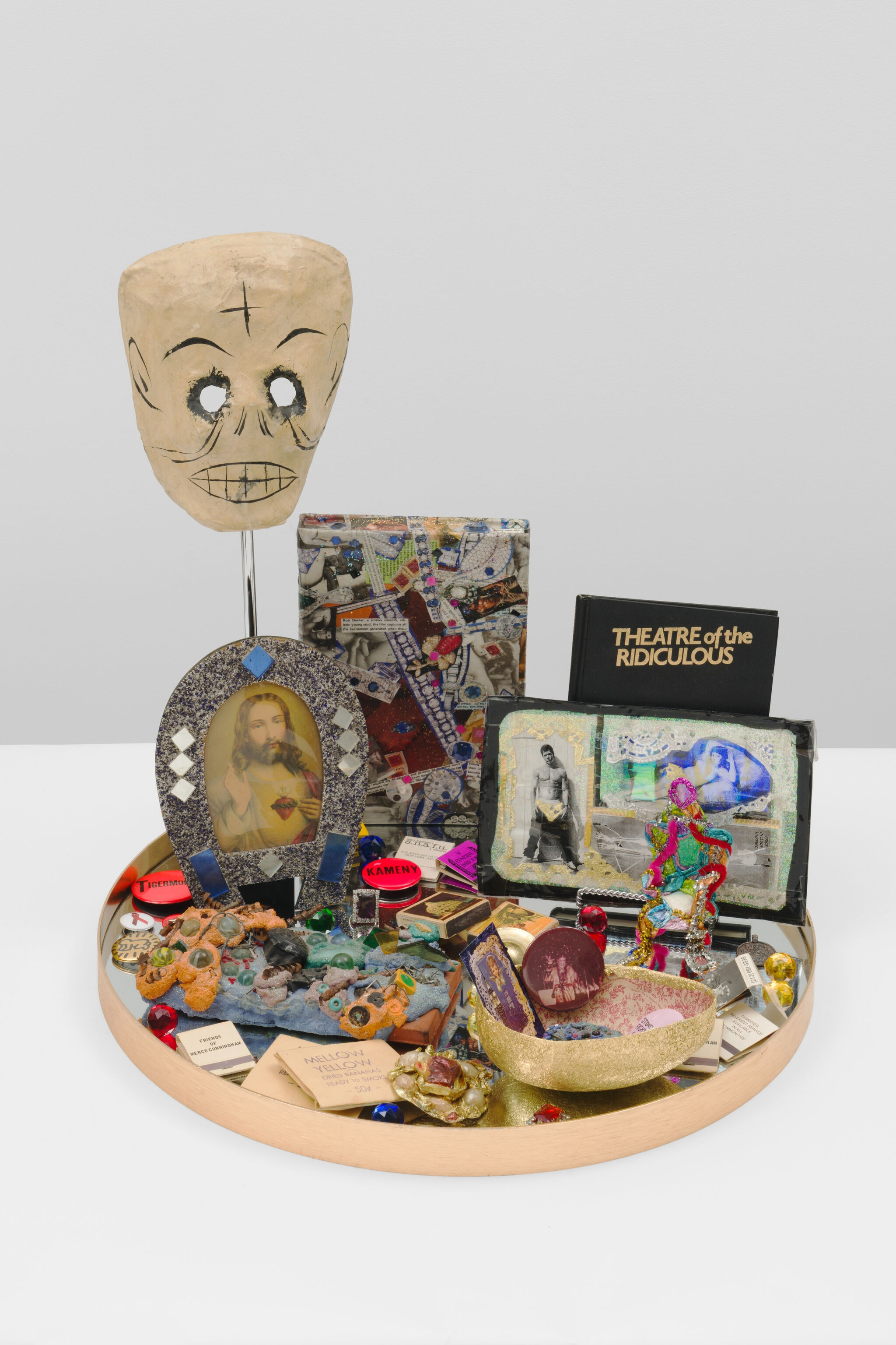 A round tray displays a papier-mâché mask, devotional image, mixed trinkets, and a small book titled Theatre of the Ridiculous.