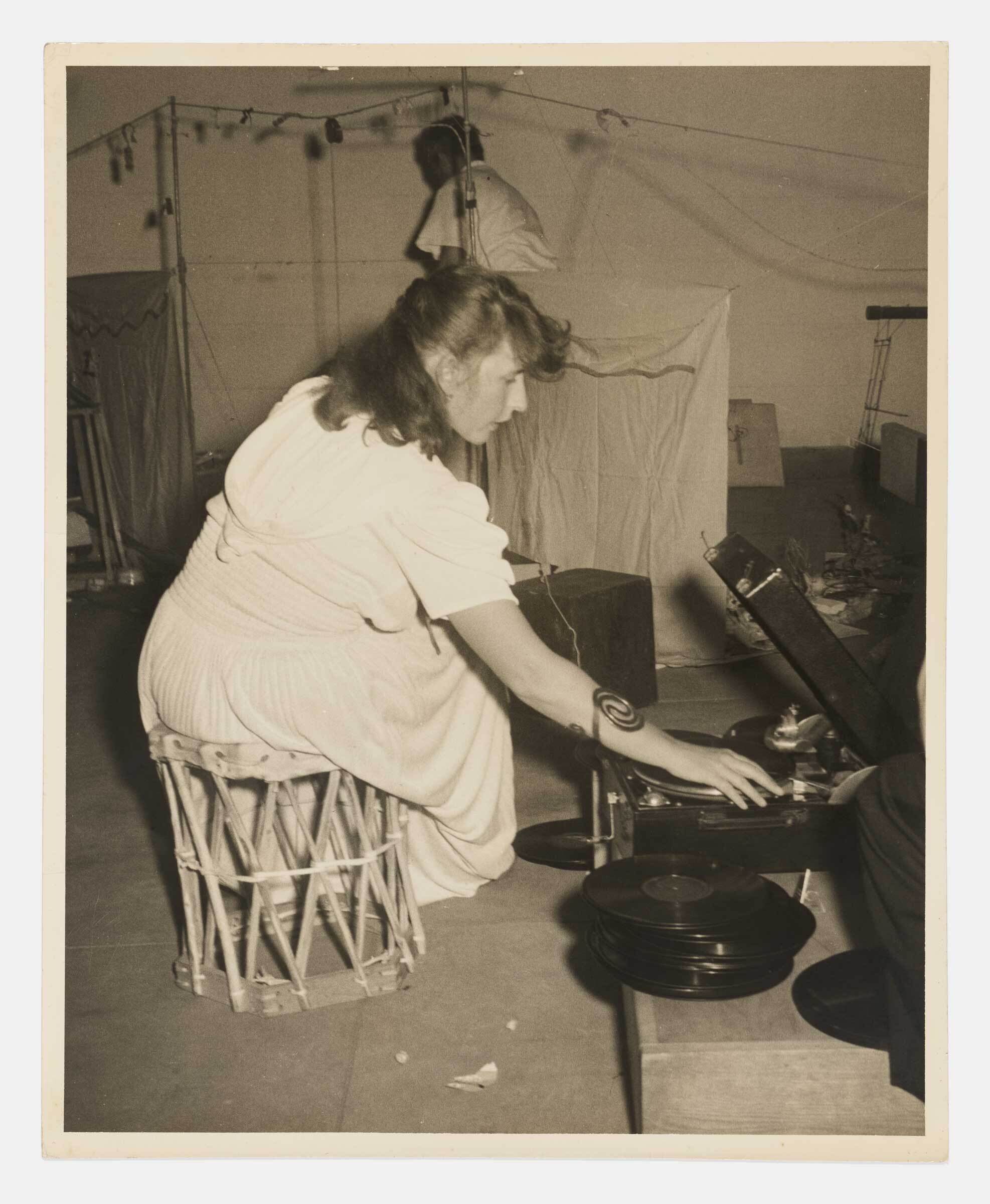A woman sits on a wicker stool and selects a record from a portable gramophone.