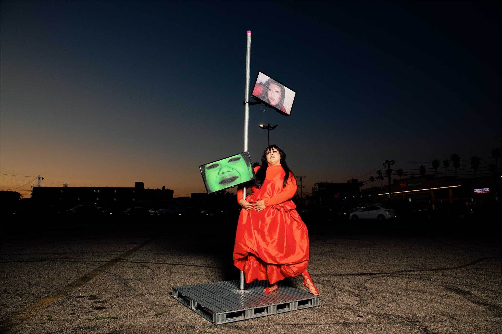 A person in a red dress stands on a platform holding a pole with two screens showing faces.