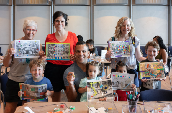 A smiling group of adults and children proudly hold up colorful mixed-media artworks at a craft table.