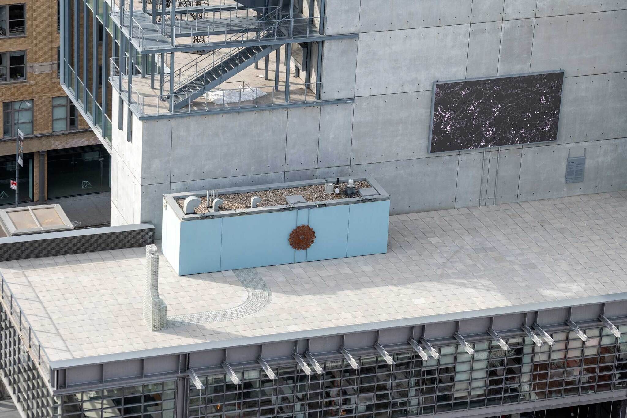 Rooftop plaza with a blue utility enclosure, decorative medallion, and a large dark display on the concrete wall.