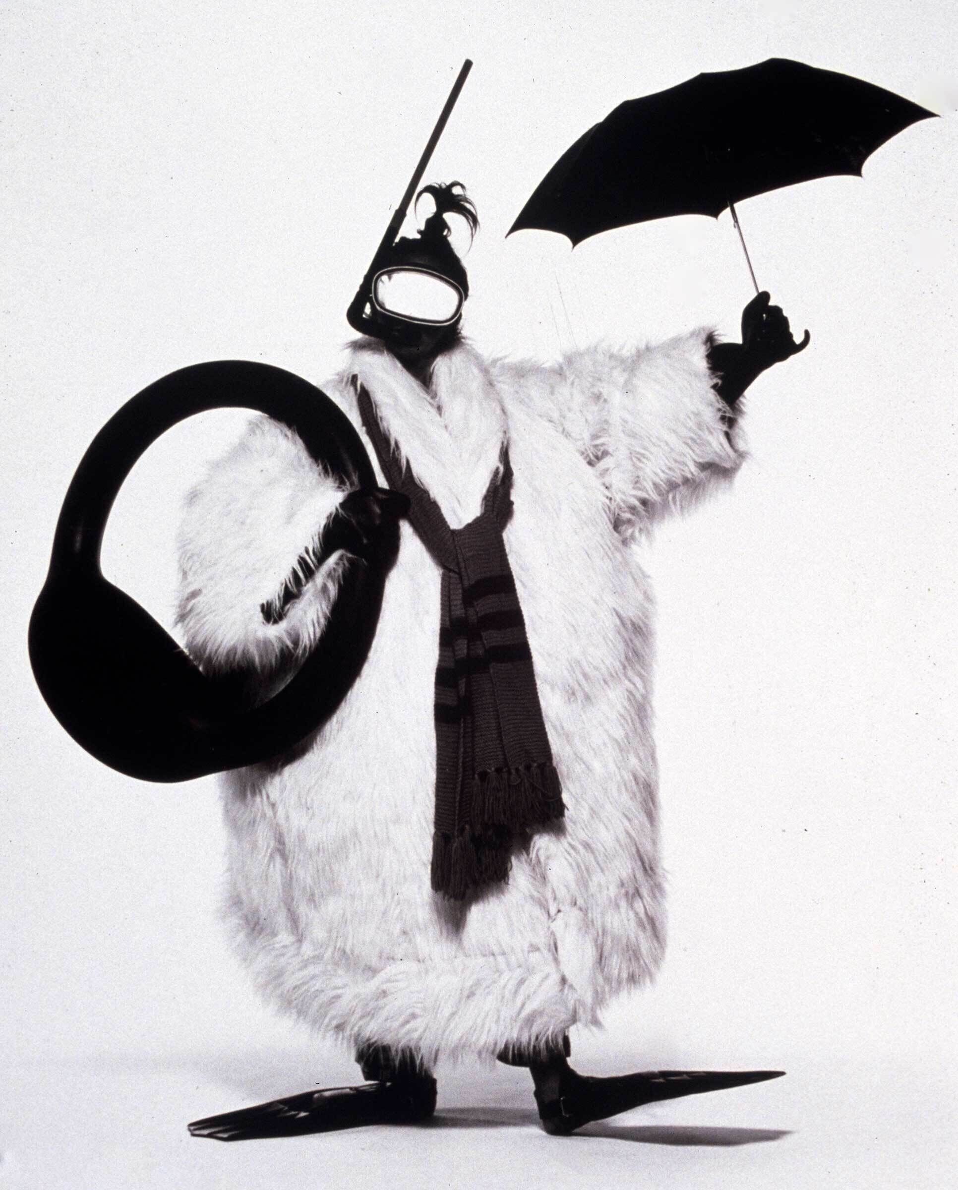 A person dressed in a fluffy coat, snorkel mask, and flippers holds an umbrella and inner tube.