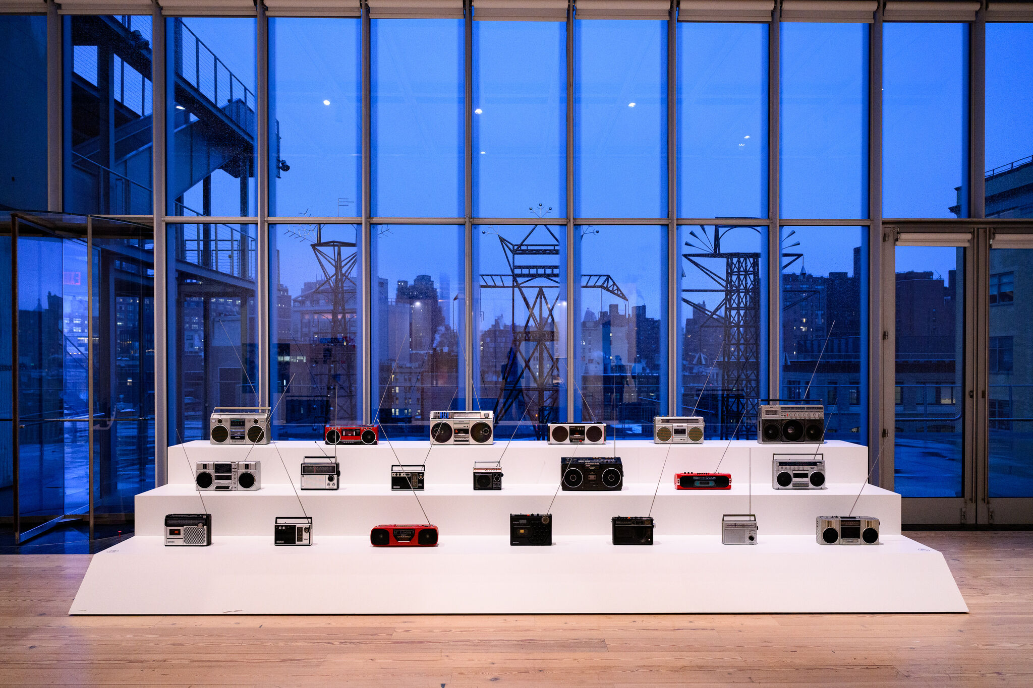Several vintage radios and boomboxes displayed on white stepped platforms in a gallery by tall windows.