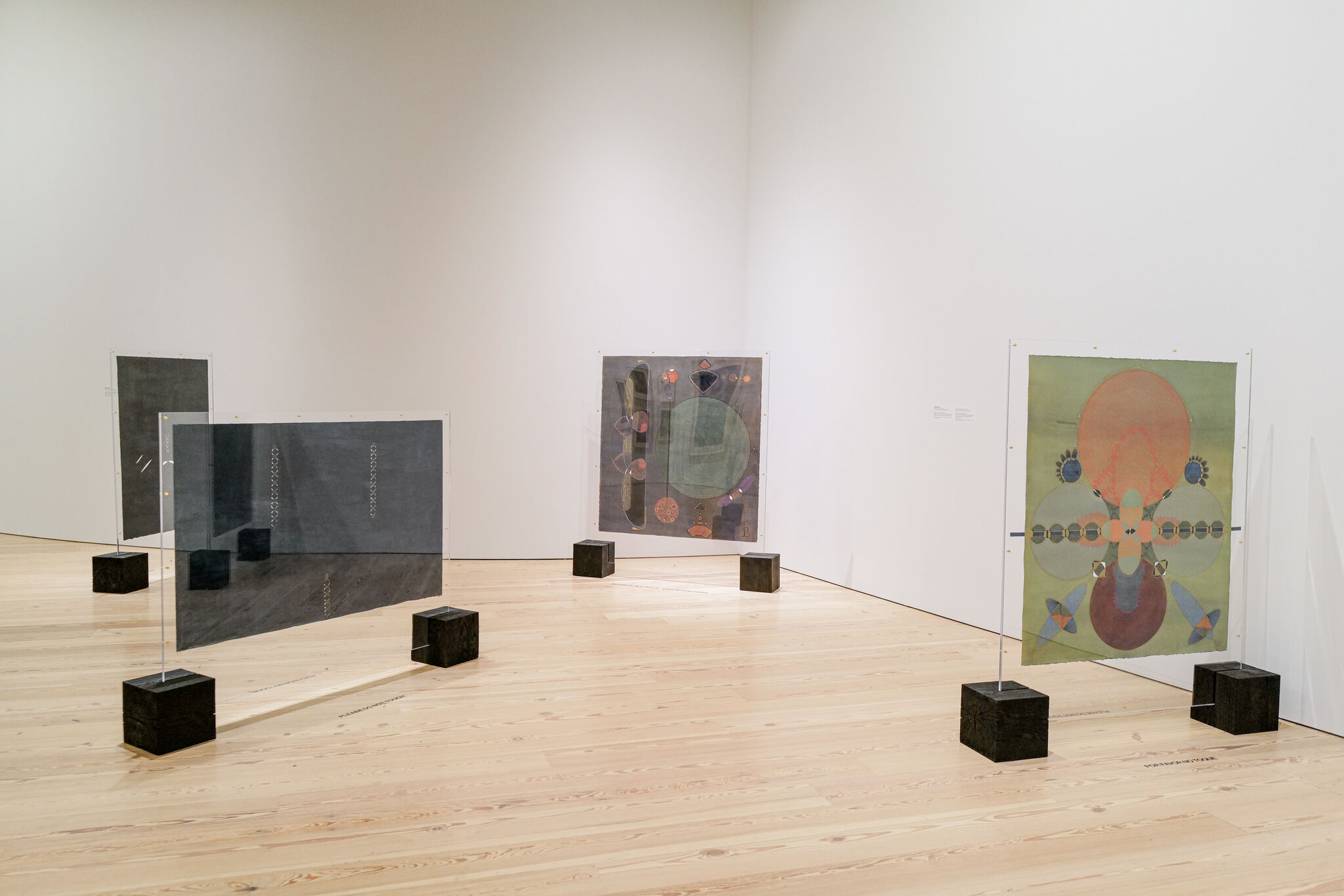 Several translucent panels with abstract geometric paintings stand on black cube bases in a white gallery.
