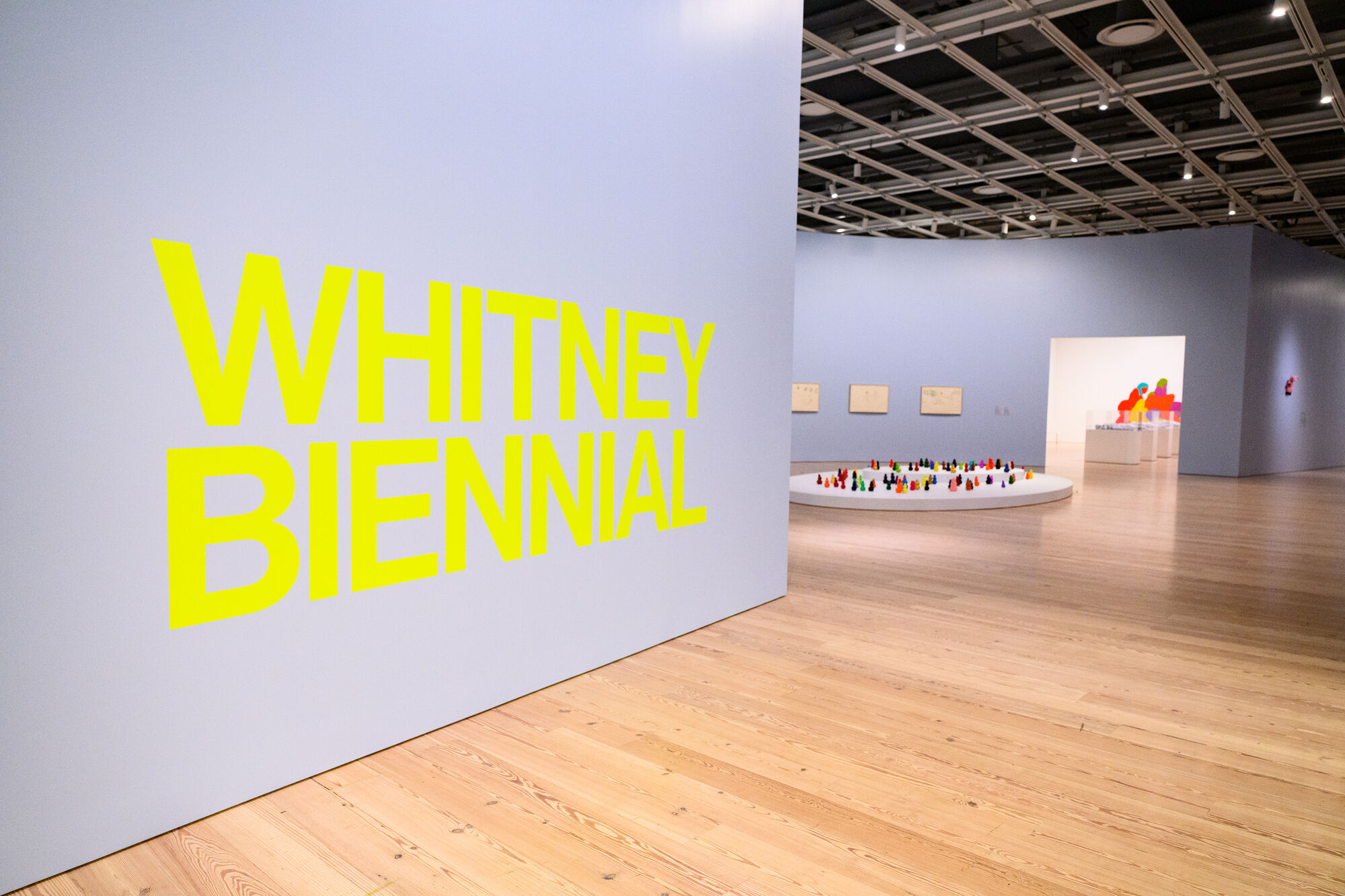 Large yellow "Whitney Biennial" lettering on a gallery wall beside a circular platform of colorful miniature sculptures.