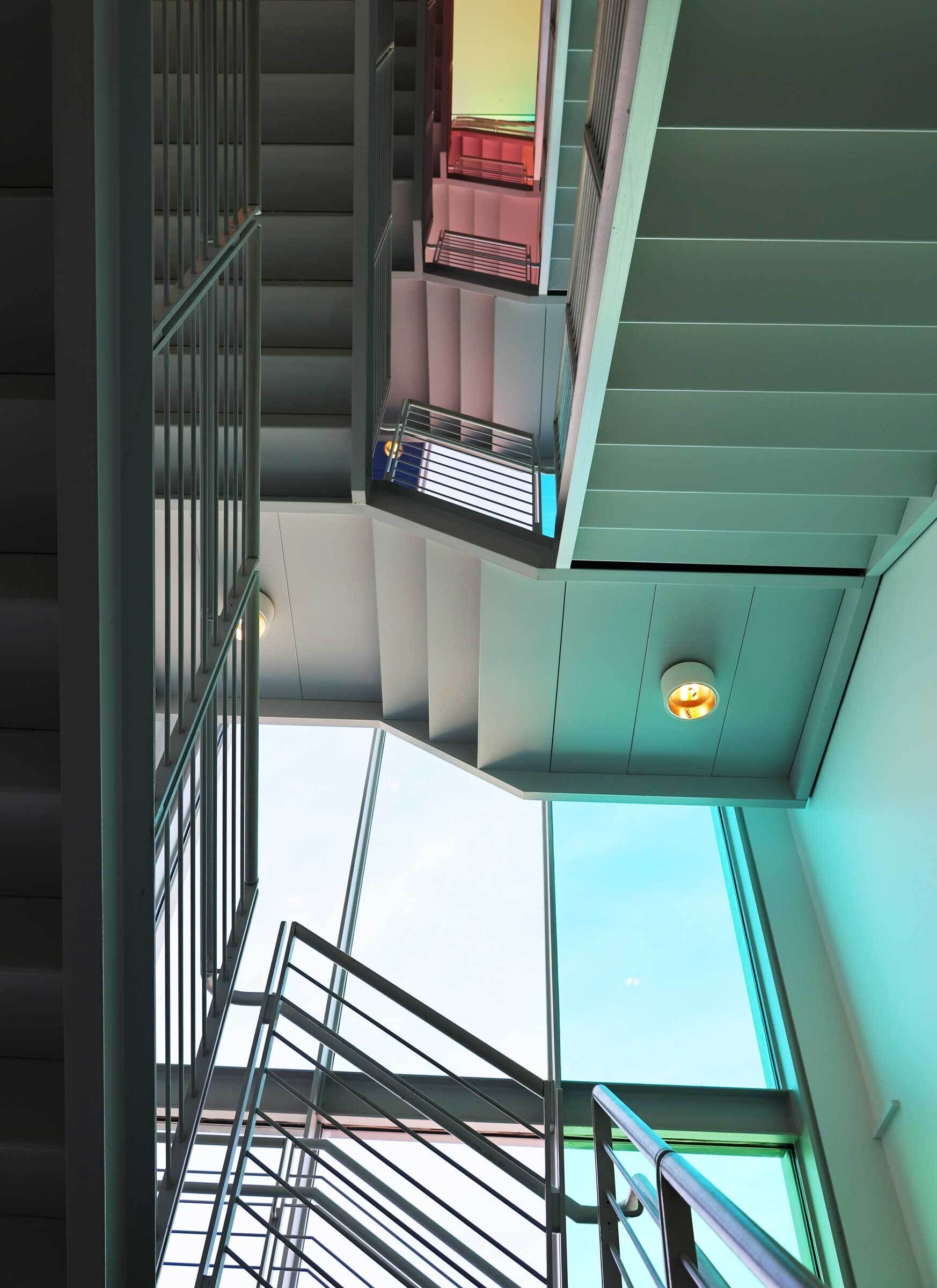 Modern stairwell with metal railings and large windows illuminated by teal and pink lighting.