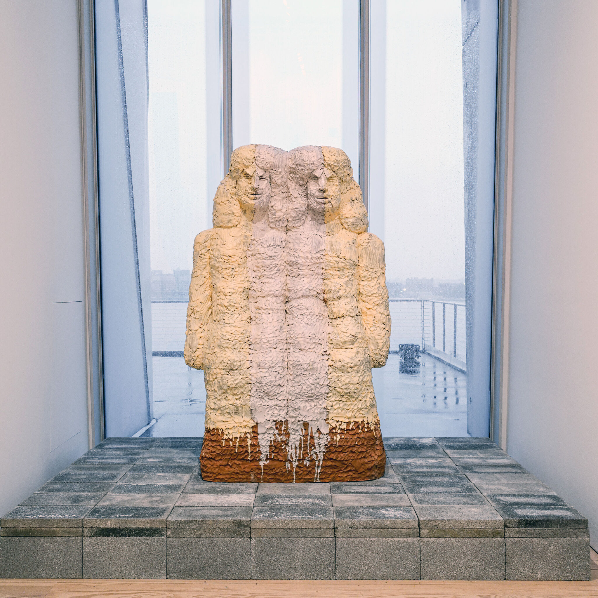 A textured three-faced sculptural figure stands on concrete blocks in a gallery window overlooking water.
