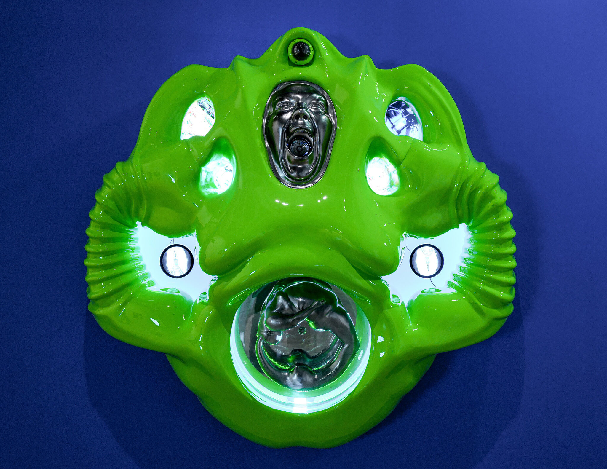 Glossy green abstract mask sculpture with metallic faces and glowing circular lights on blue.