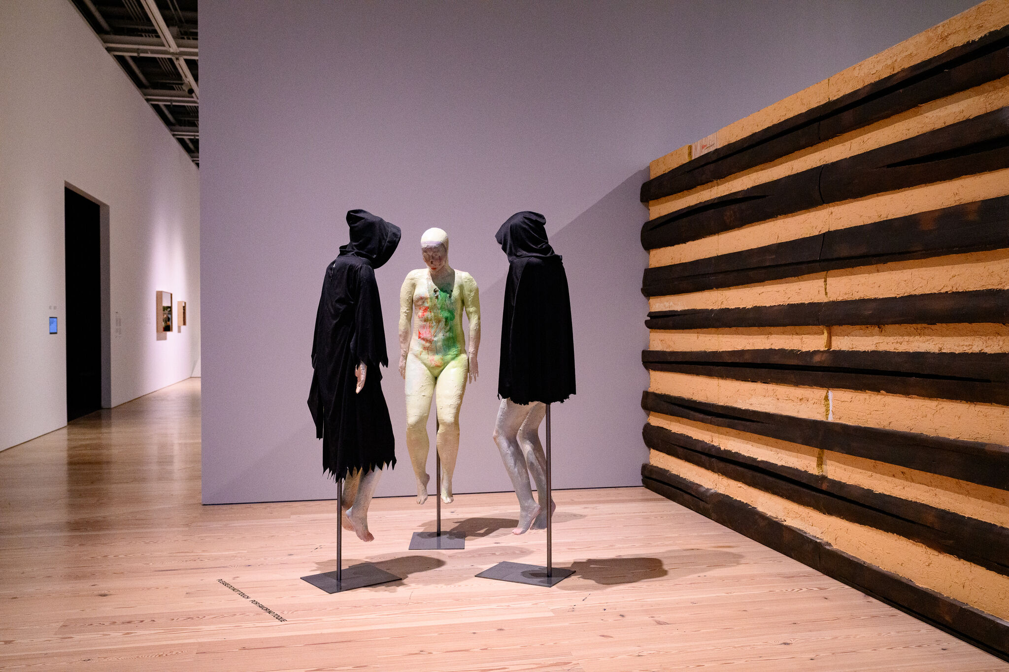 Three suspended mannequins in a gallery: a paint-covered pale figure flanked by two hooded black figures.