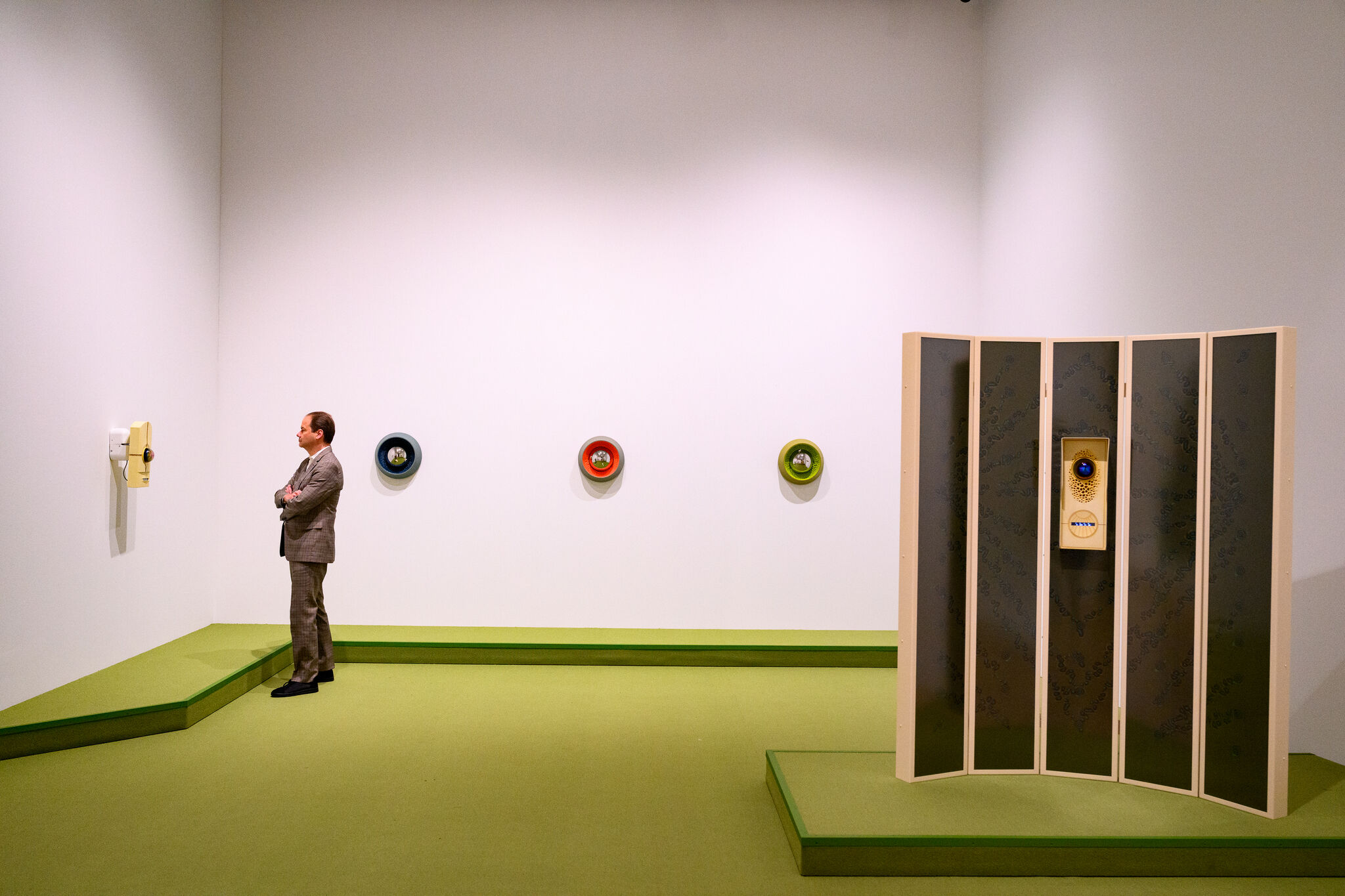 A man in a suit stands with arms crossed observing wall-mounted circular art and a yellow telephone piece.