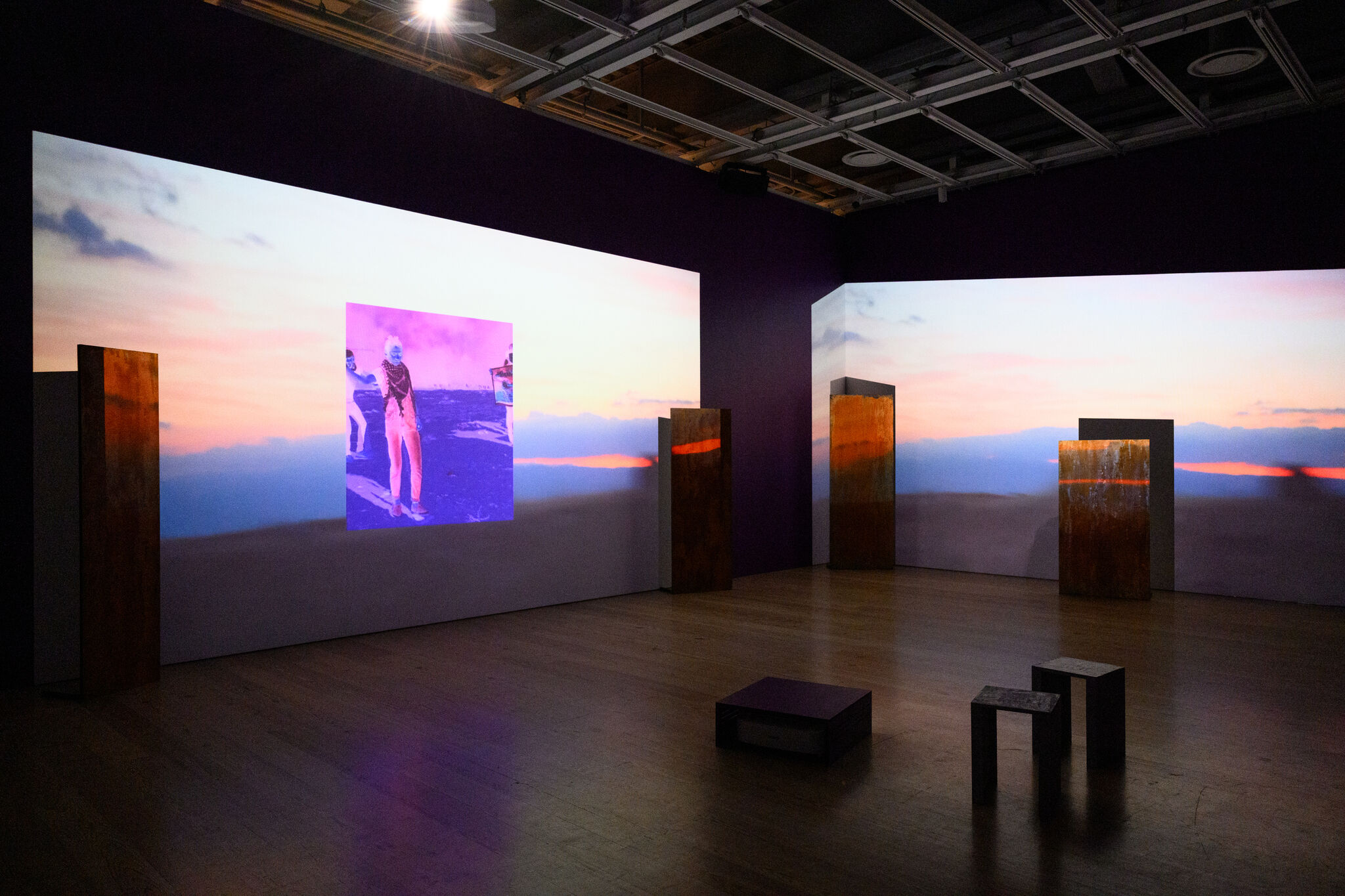 Gallery installation with wide sunset projections, rusted vertical panels, and small stools on the wooden floor.