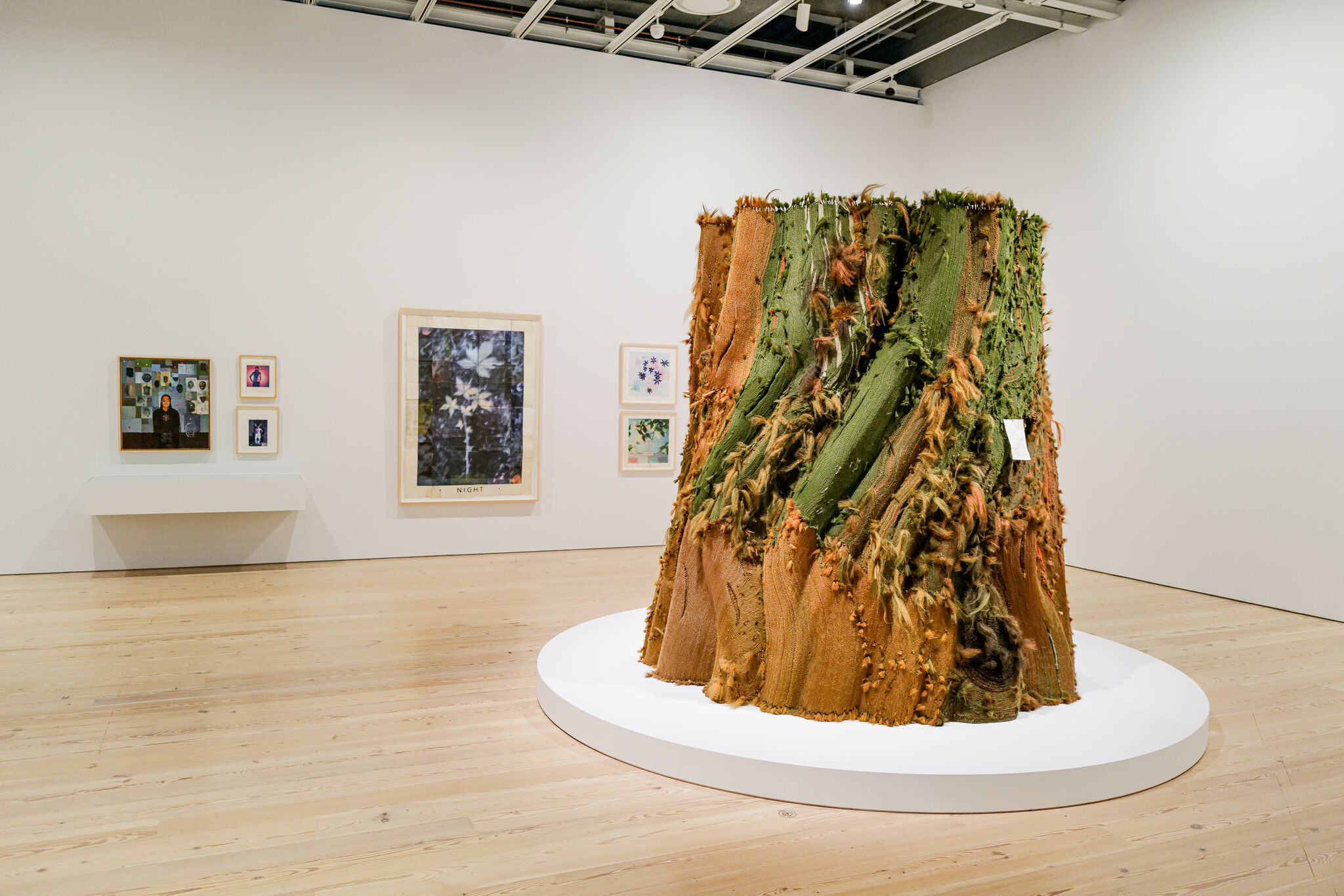 A large textured green and orange textile sculpture stands on a white circular platform in a gallery.