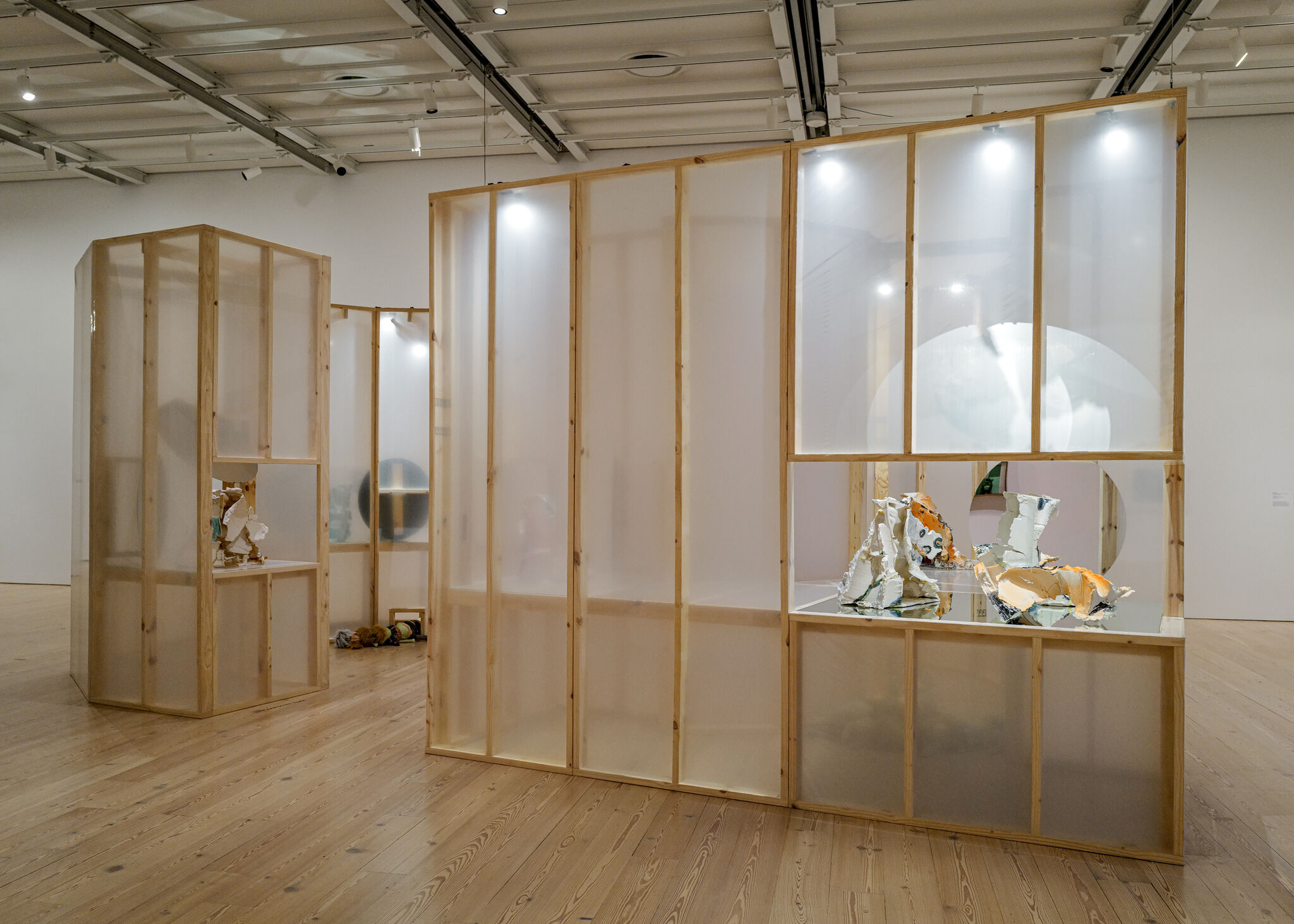 Wooden-framed translucent panels form partitioned gallery spaces displaying fragmented ceramic sculptures on mirrored surfaces.