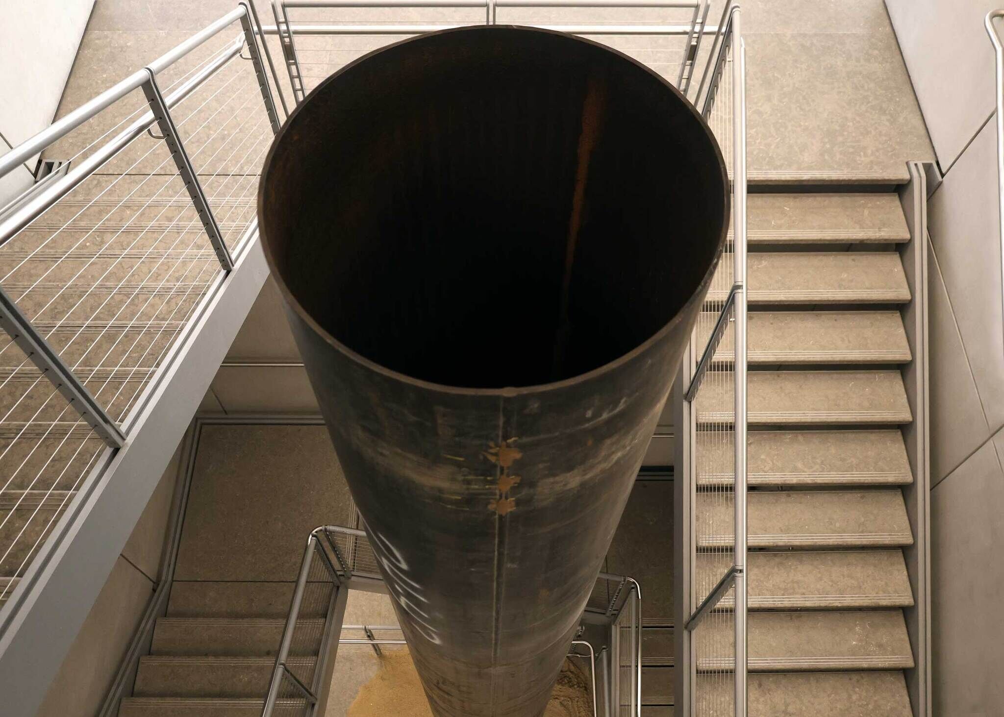 A large hollow metal column stands vertically through a stairwell, seen from above.
