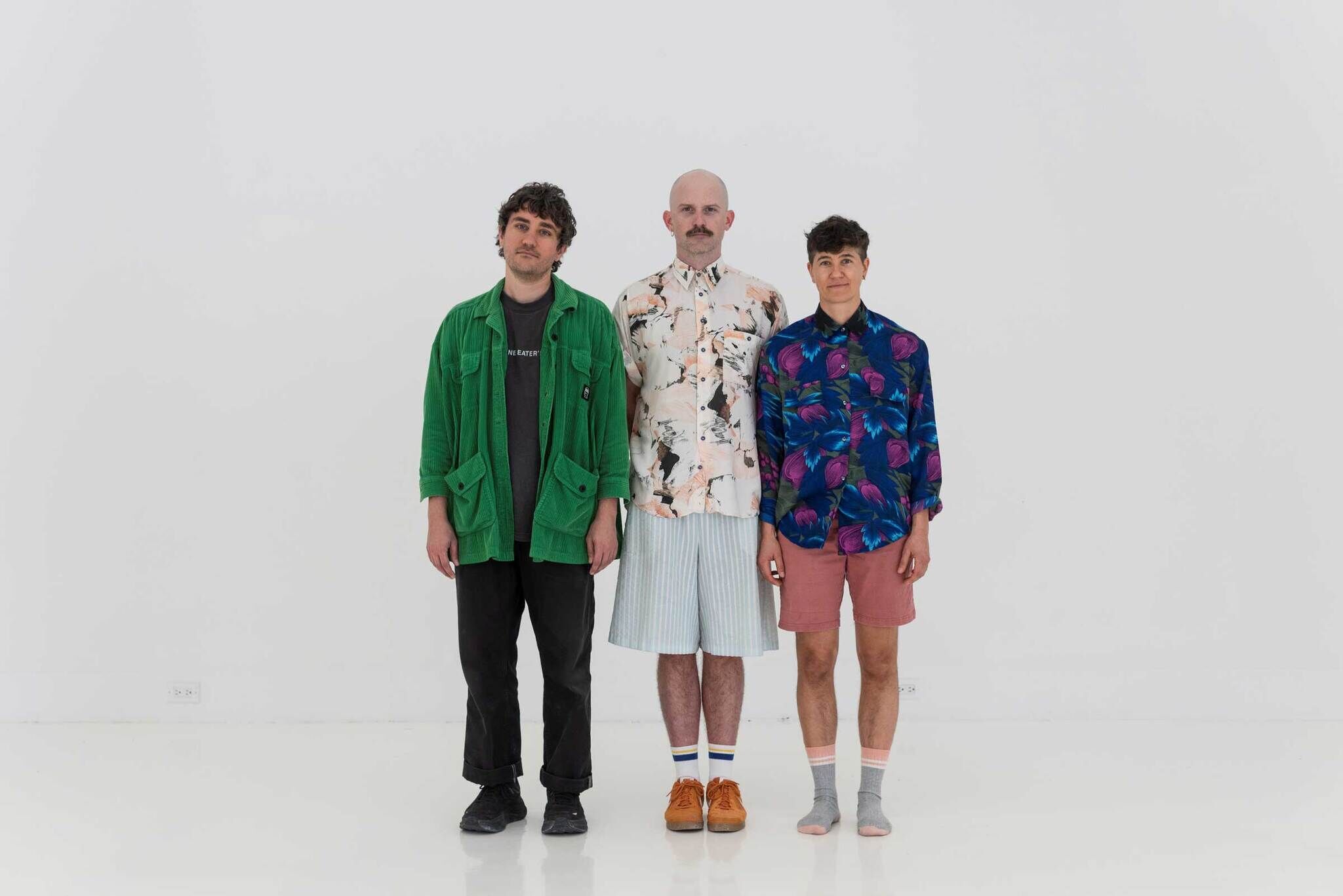 Three people stand side by side against a white wall wearing colorful patterned shirts and shorts.